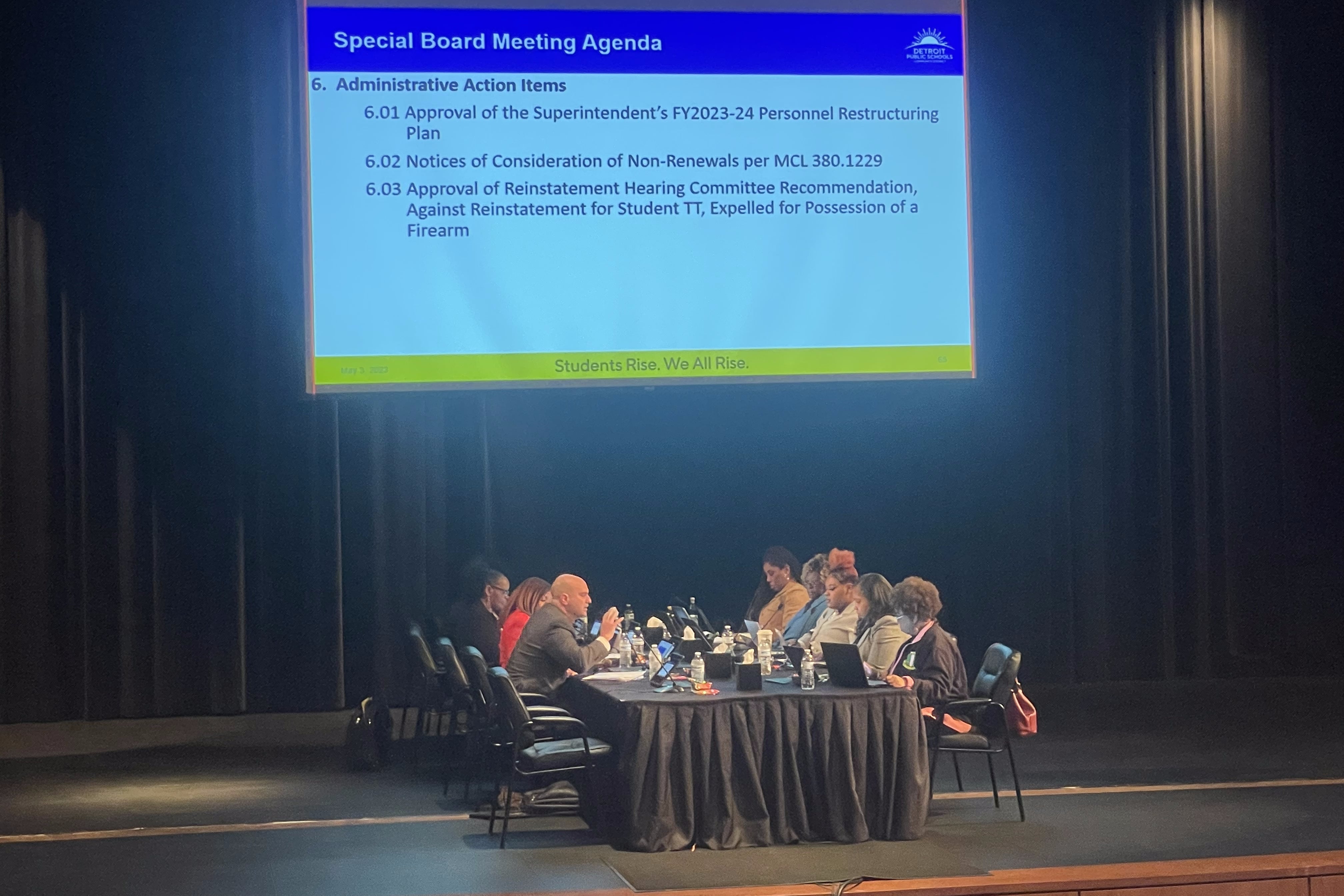 A board of people seated at a table on a stage in front of a projection screen showing text from a slide show.