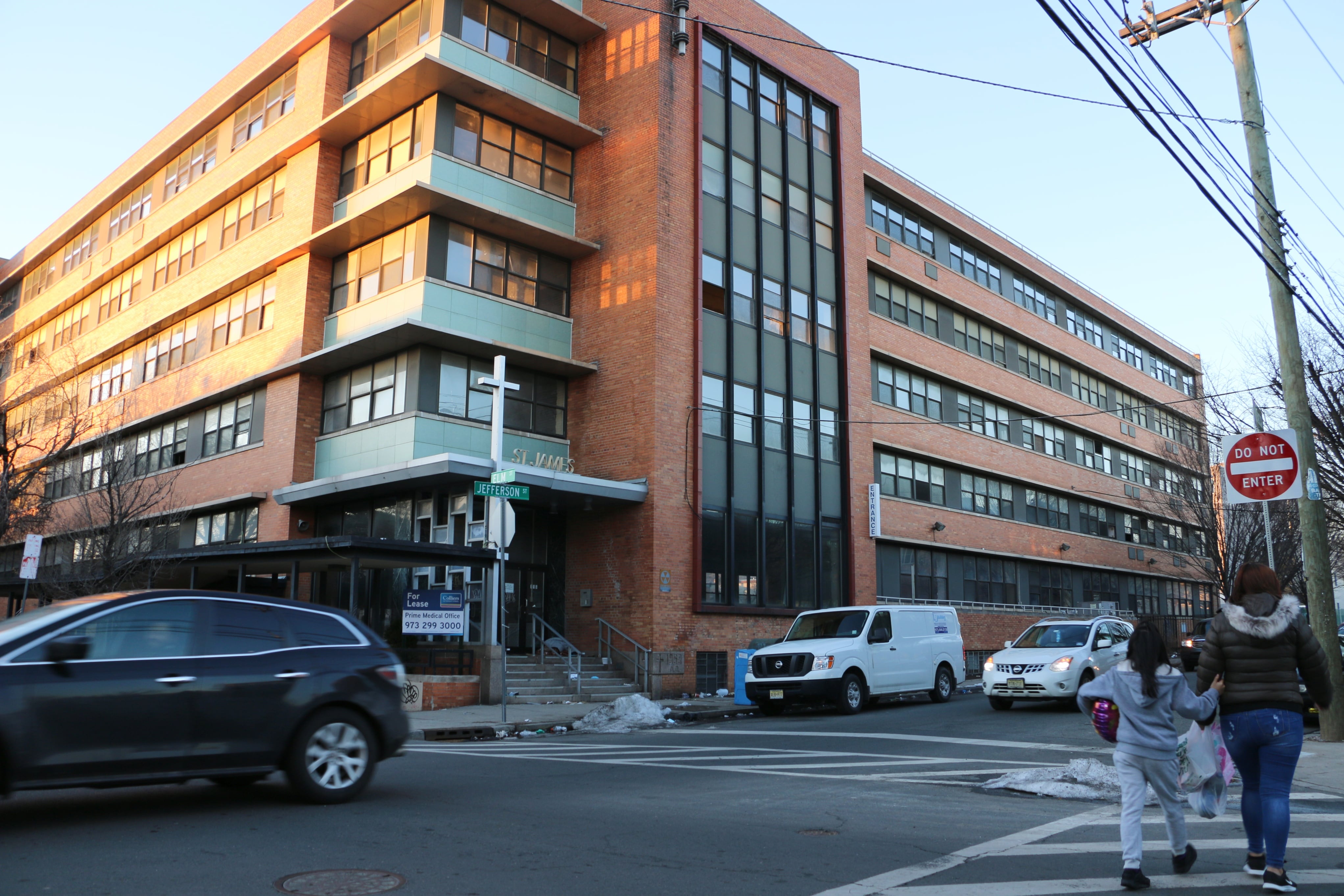 Two people cross an intersection while a car drives by them in front of a large brick school building.