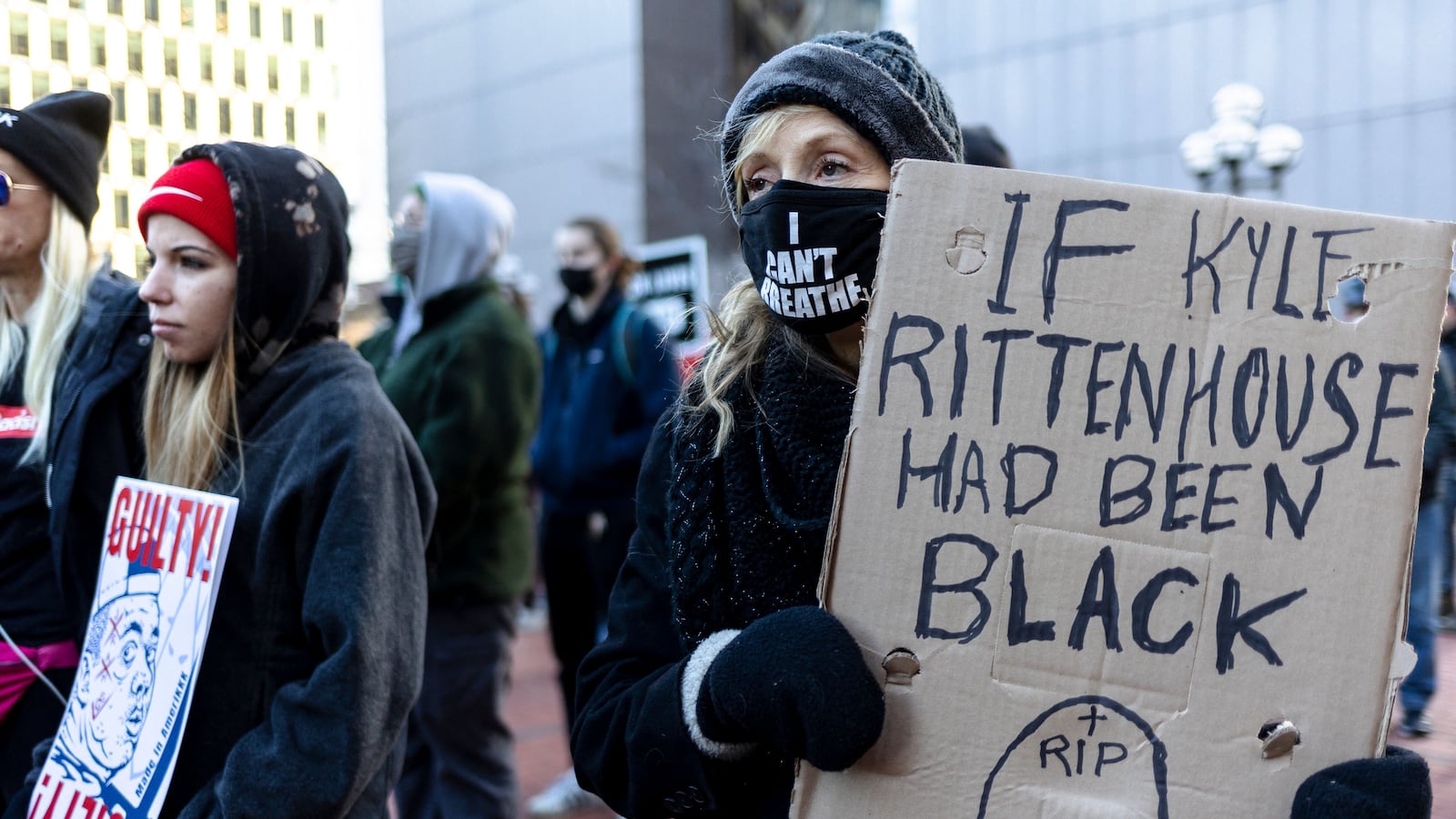 At a protest, a woman holds a sign that reads, “If Kyle Rittenhouse had been black,” with an illustration of a headstone that reads “RIP” at the top. She is also wearing a mask that reads, “I Can’t Breathe”.