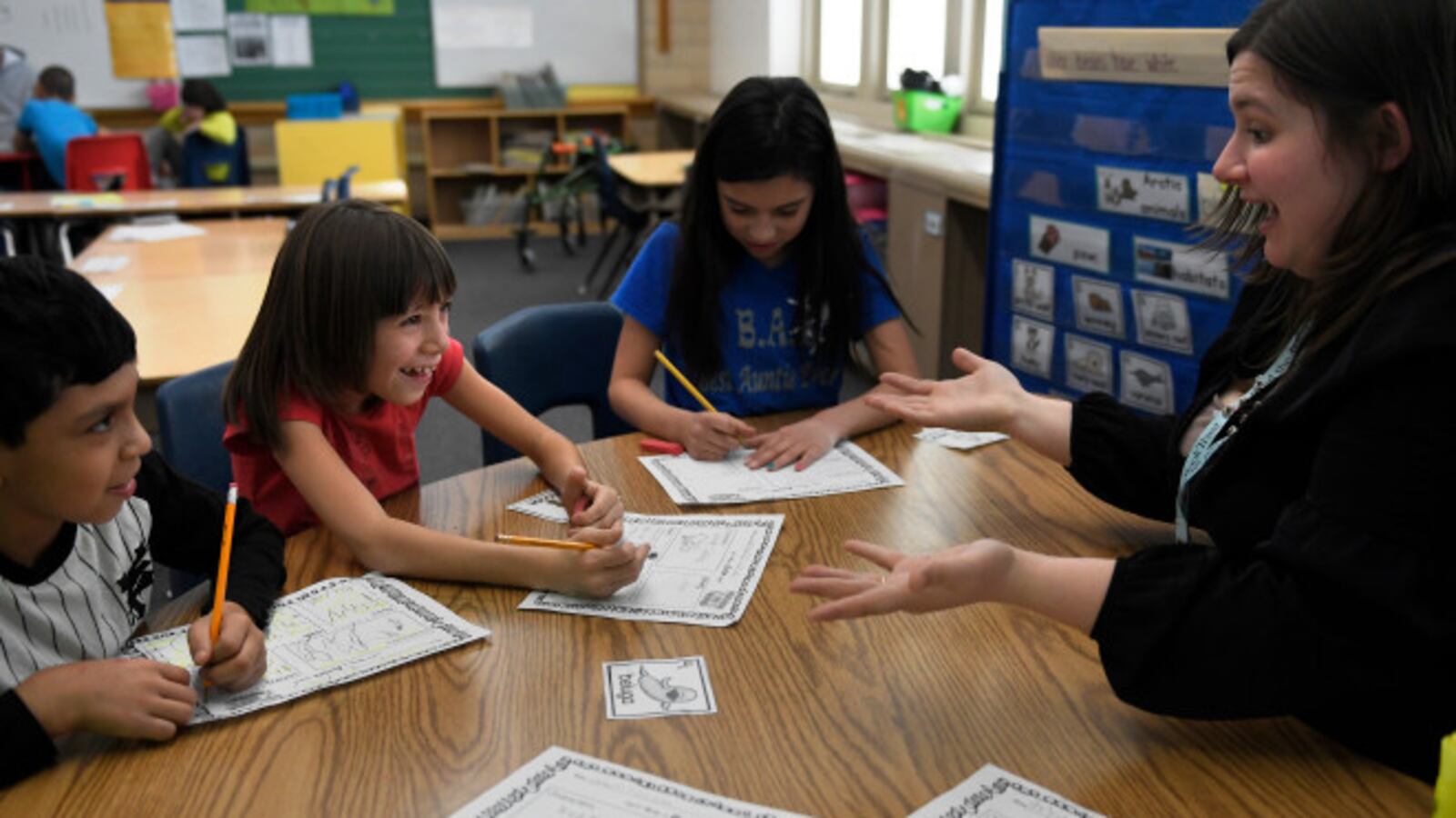 A teacher and three elementary school students sit at a table in a classroom. The teacher has a surprised look on her face, and one of the students is laughing.