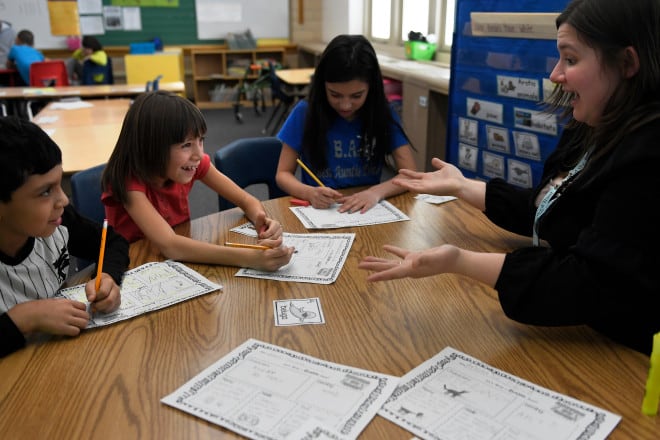 A teacher and three elementary school students sit at a table in a classroom. The teacher has a surprised look on her face, and one of the students is laughing.