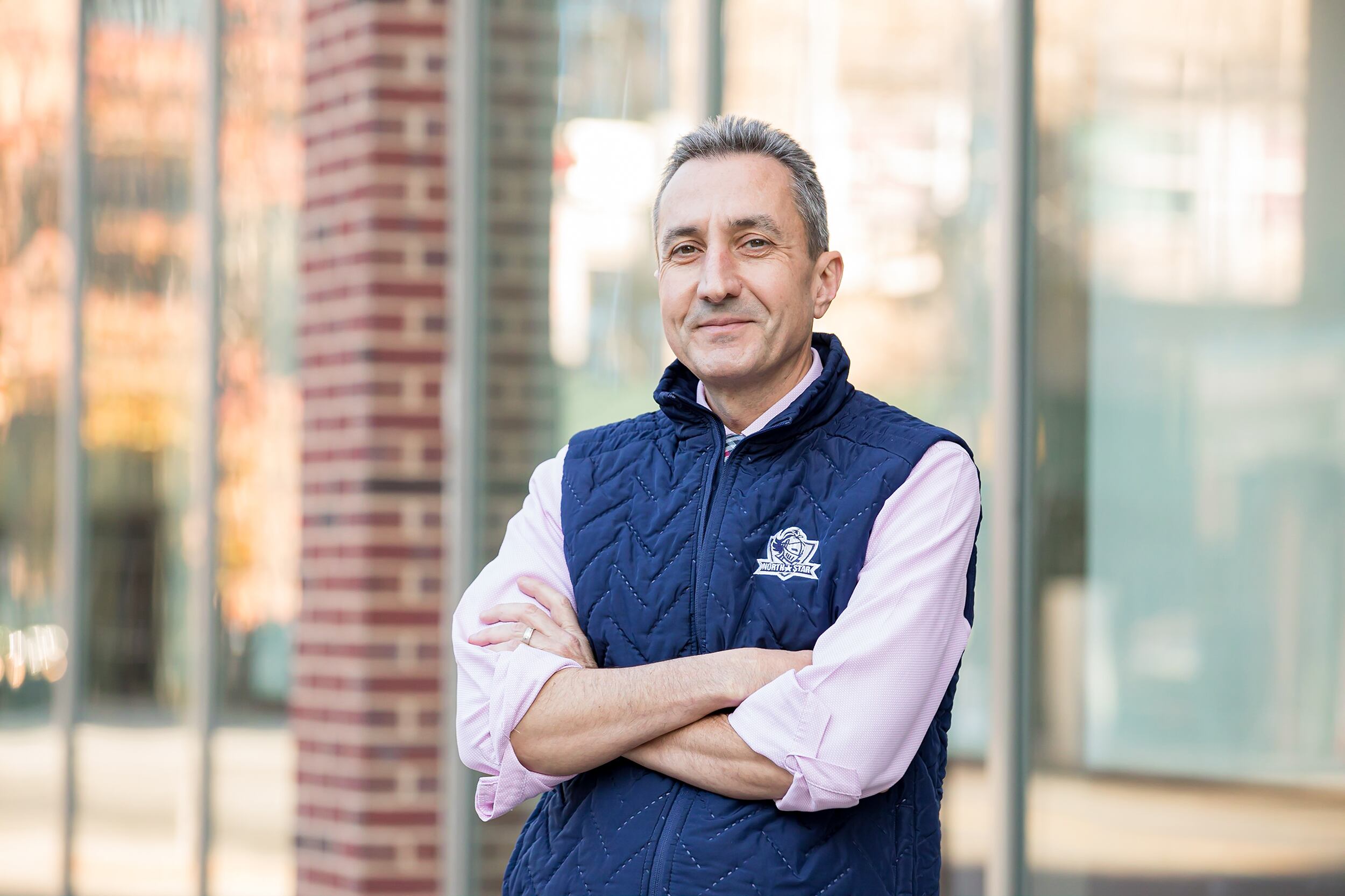 A photograph of a man in a blue vest and pink dress shirt poses for a photo in front of a building.