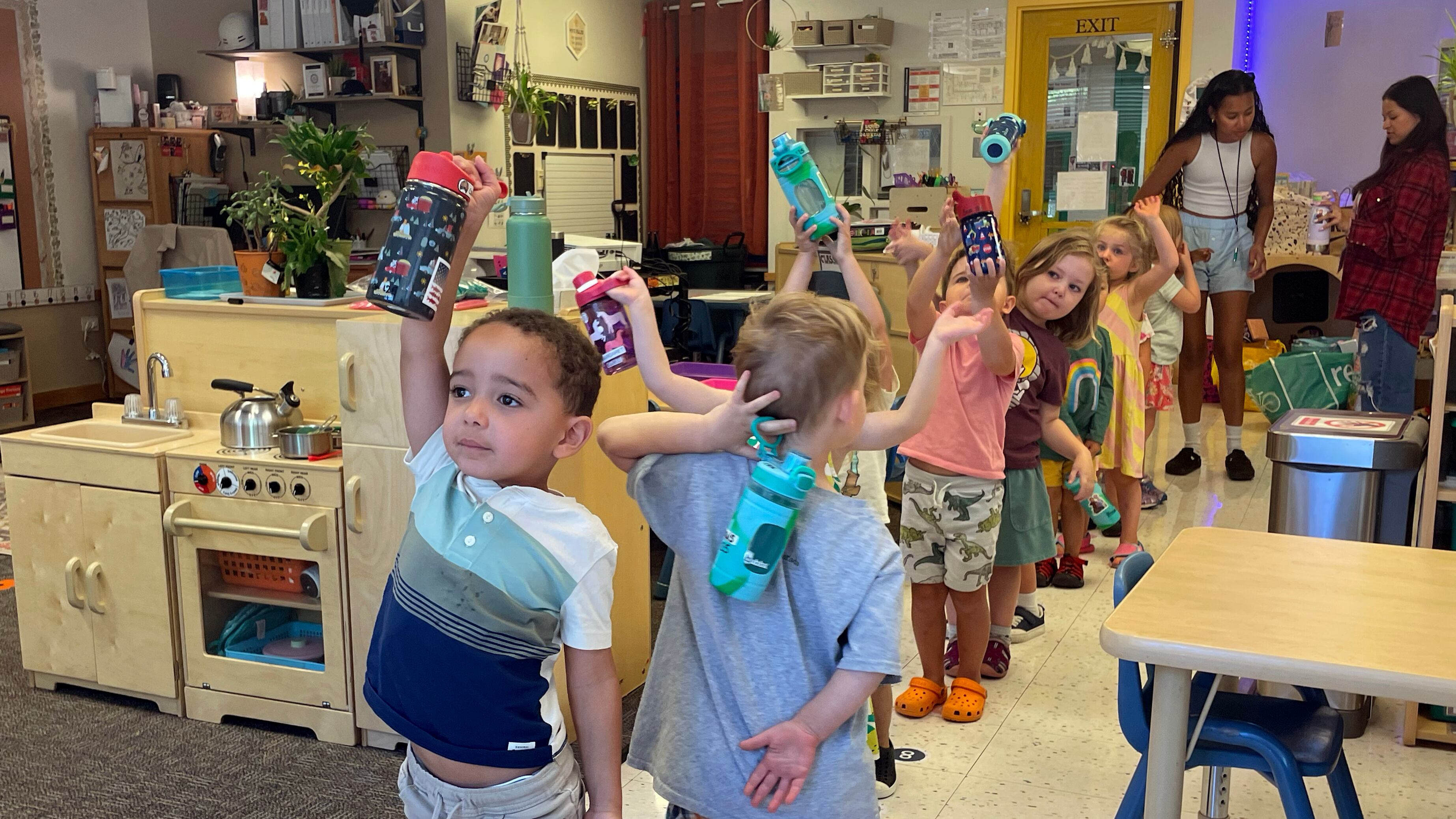 Preschool students in a line hold water bottles over their heads.