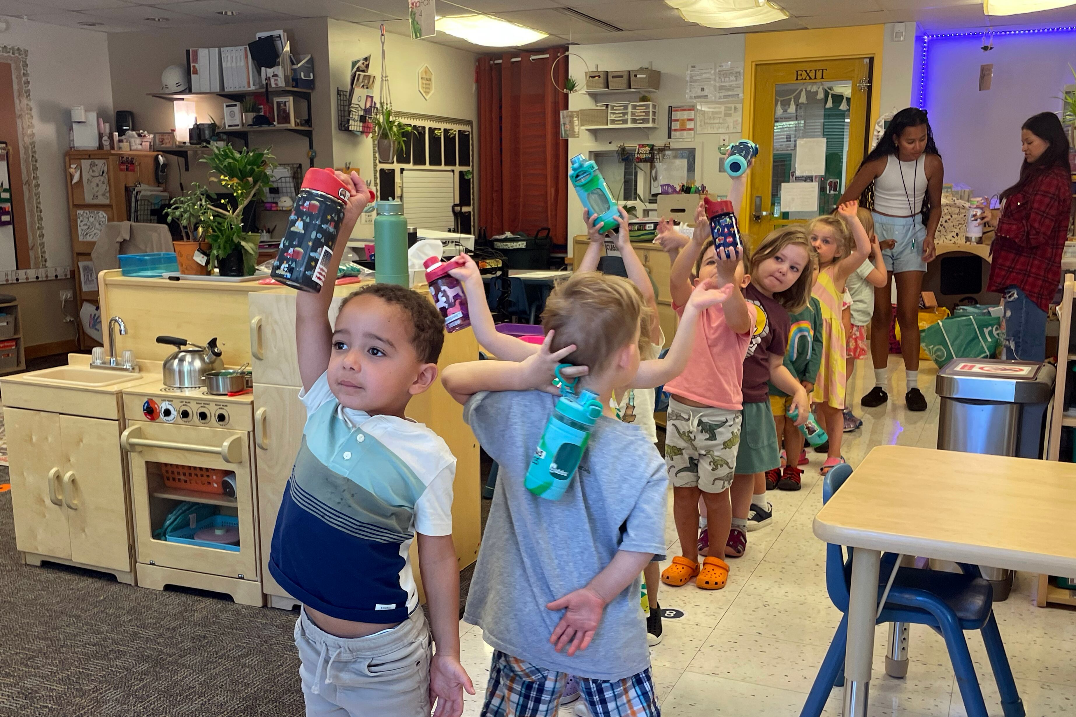 Preschool students in a line hold water bottles over their heads.