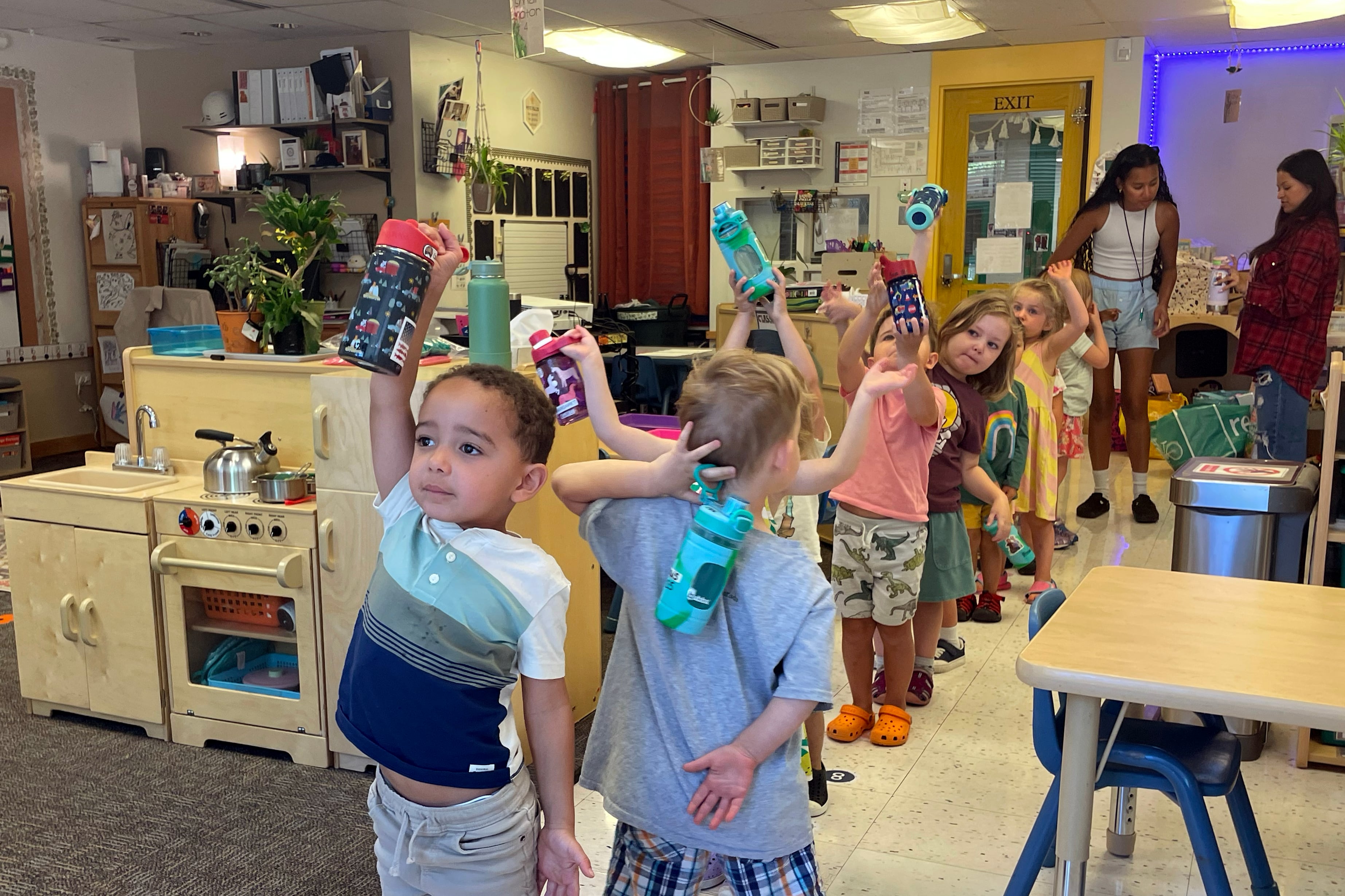 Preschool students in a line hold water bottles over their heads.