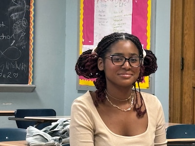 A photograph of a young Black student wearing a tan shirt and sitting at a desk in a classroom.