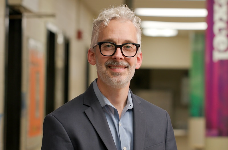 A man with white hair and black glasses smiles for a portrait in the middle of a school hallway.