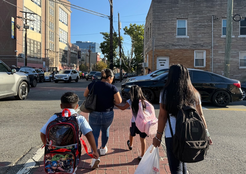 A photograph of an adult and three students wearing backpacks cross a street on a sunny day.
