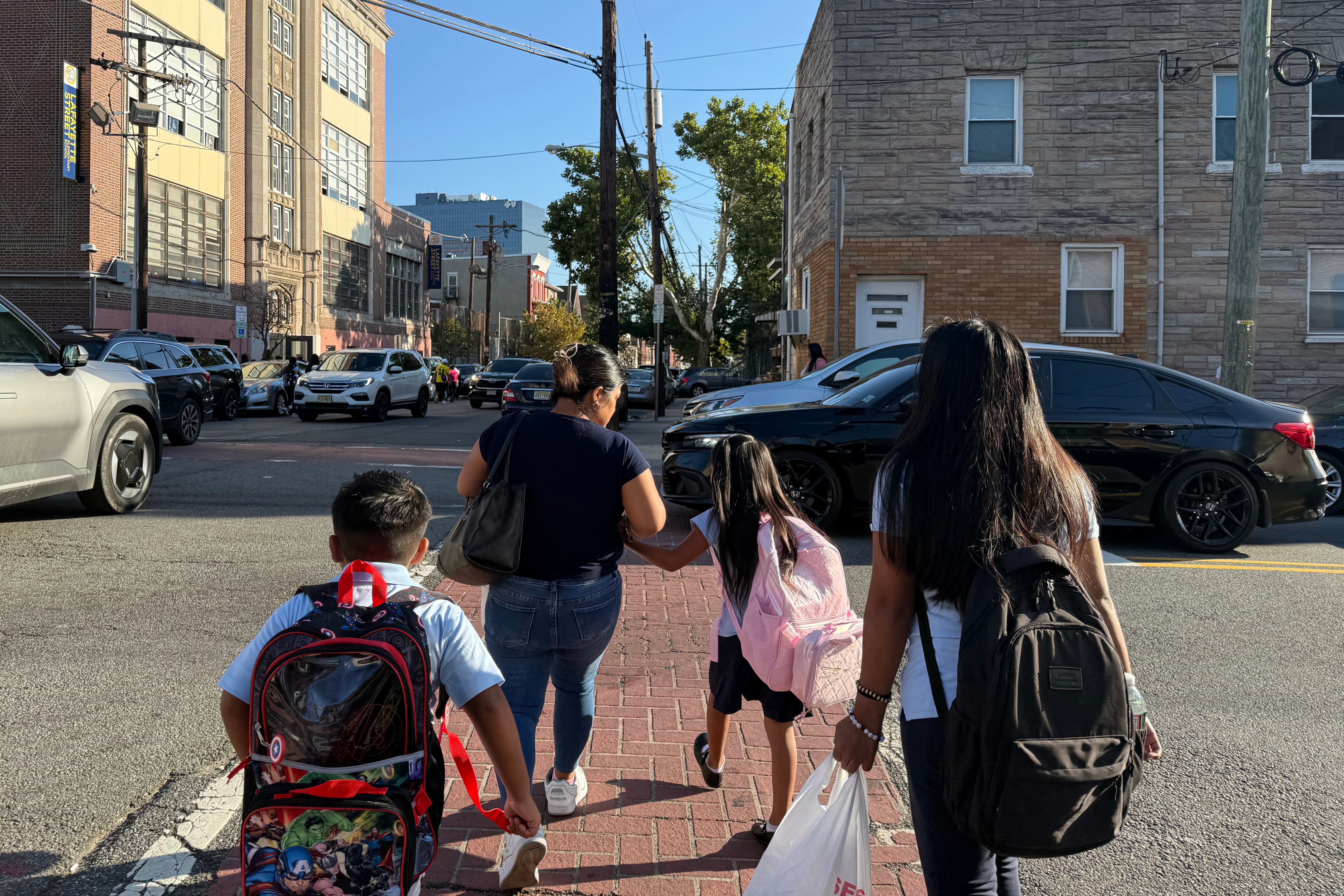 A photograph of an adult and three students wearing backpacks cross a street on a sunny day.