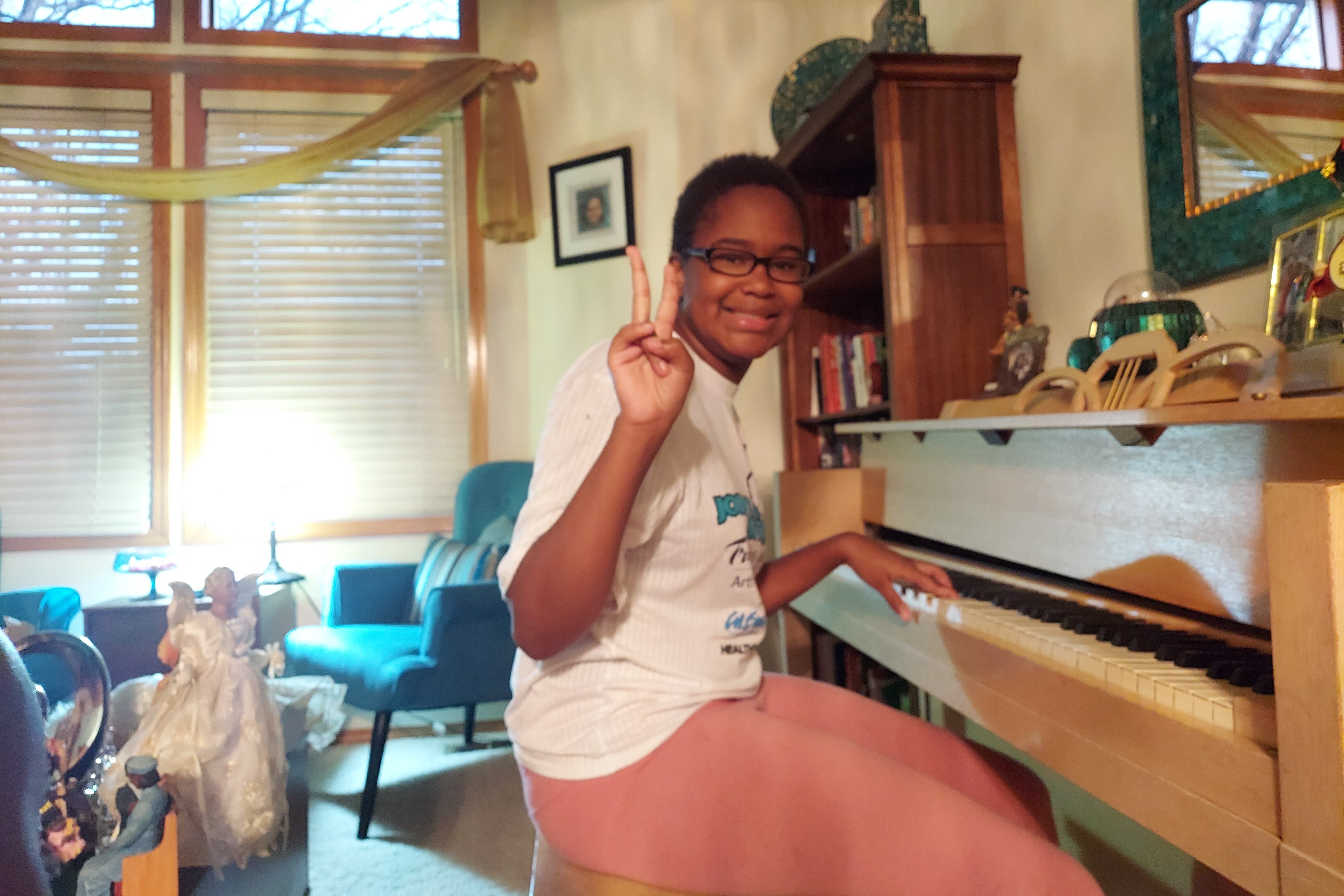 Karinne Jones, 14, sits at a piano and holds up a peace sign at the camera while smiling.