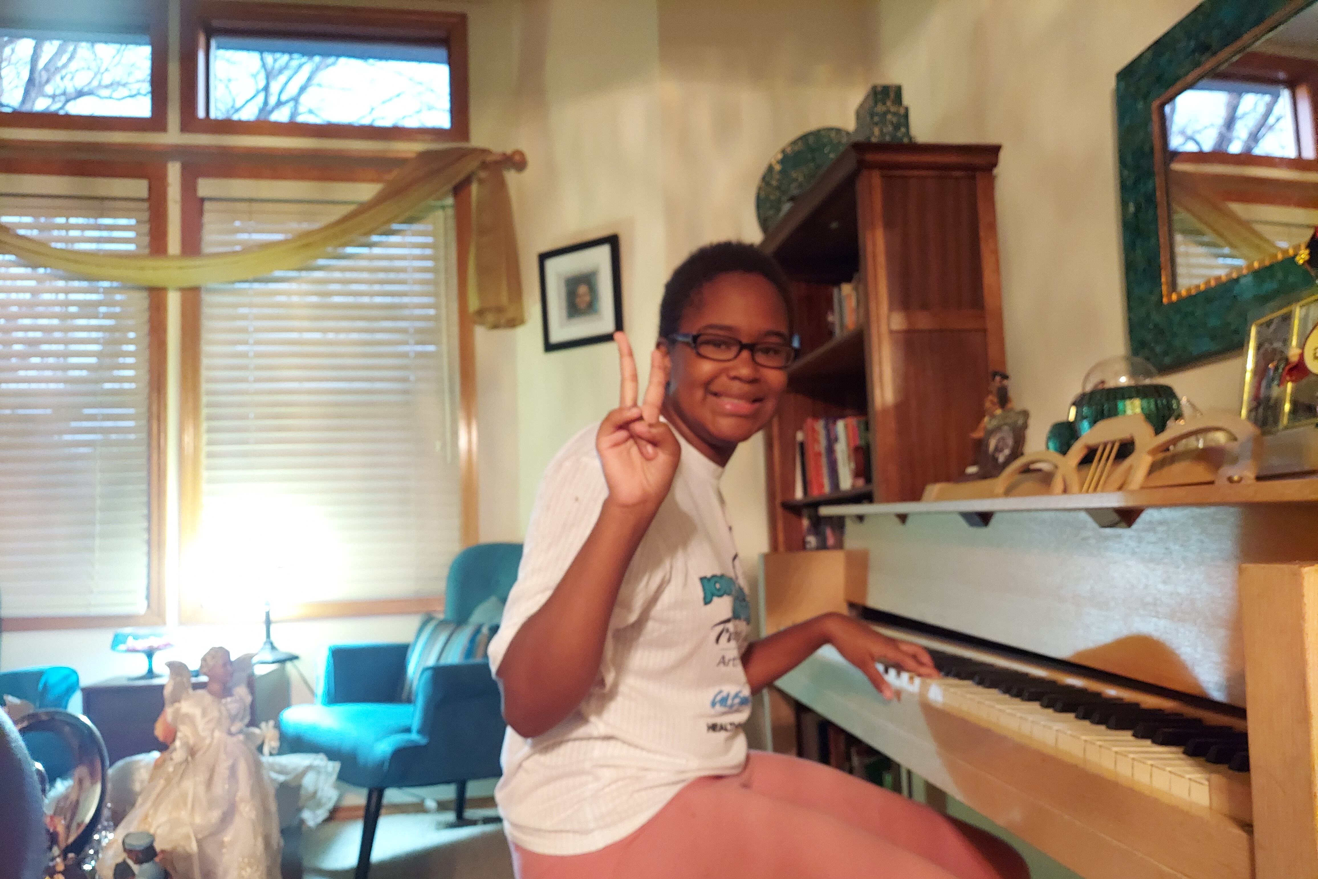 Karinne Jones, 14, sits at a piano and holds up a peace sign at the camera while smiling.