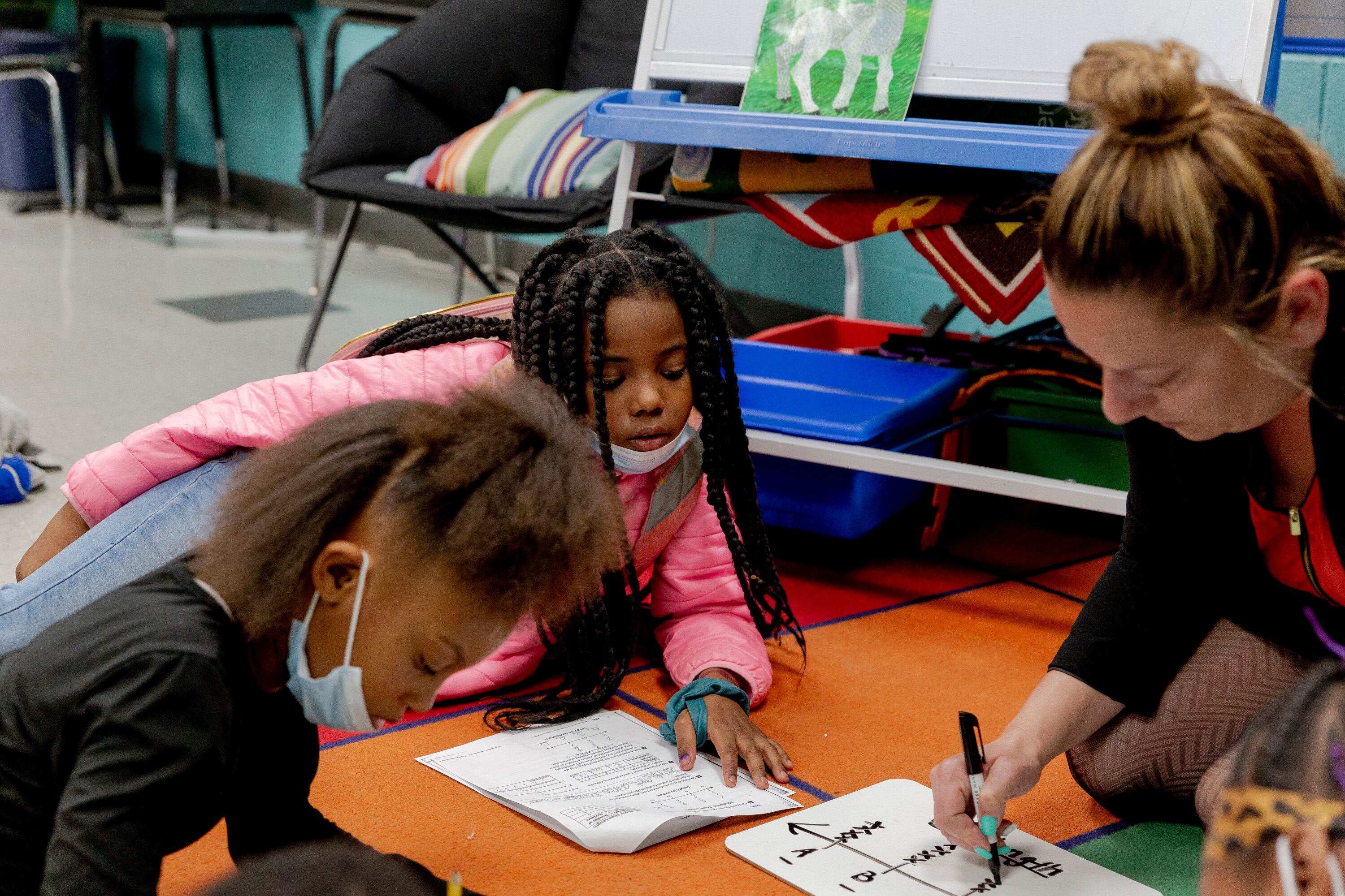 Two young girls work on a math problem while seated on a carpet as their school principal writes the solution with a black marker.
