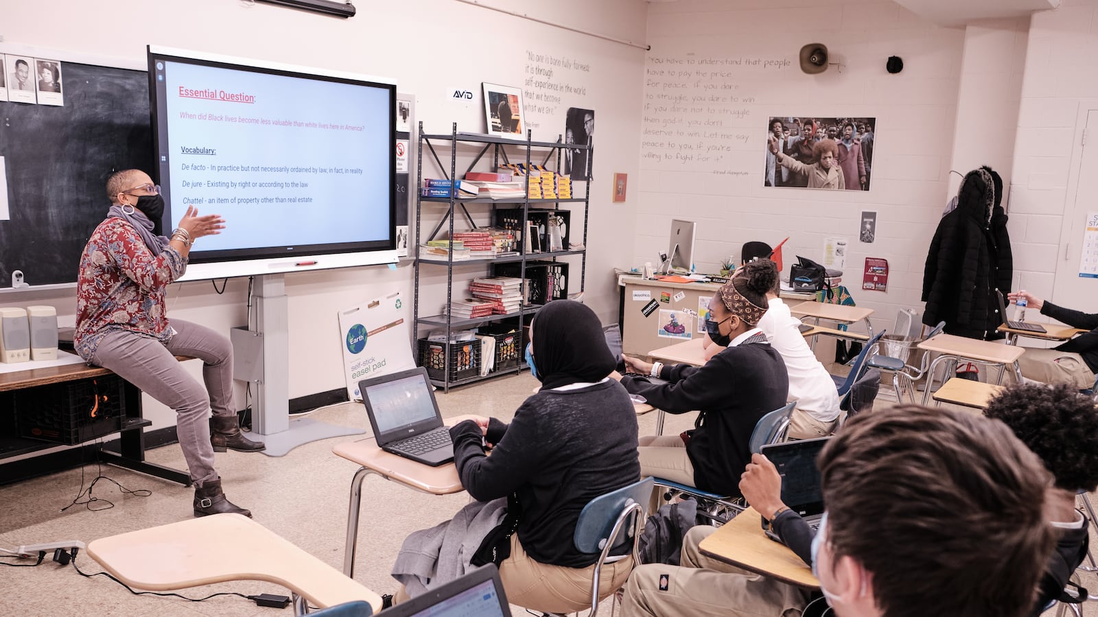 A teacher points at a screen while sitting on a desk in front of a group of students sitting at desks with laptops.