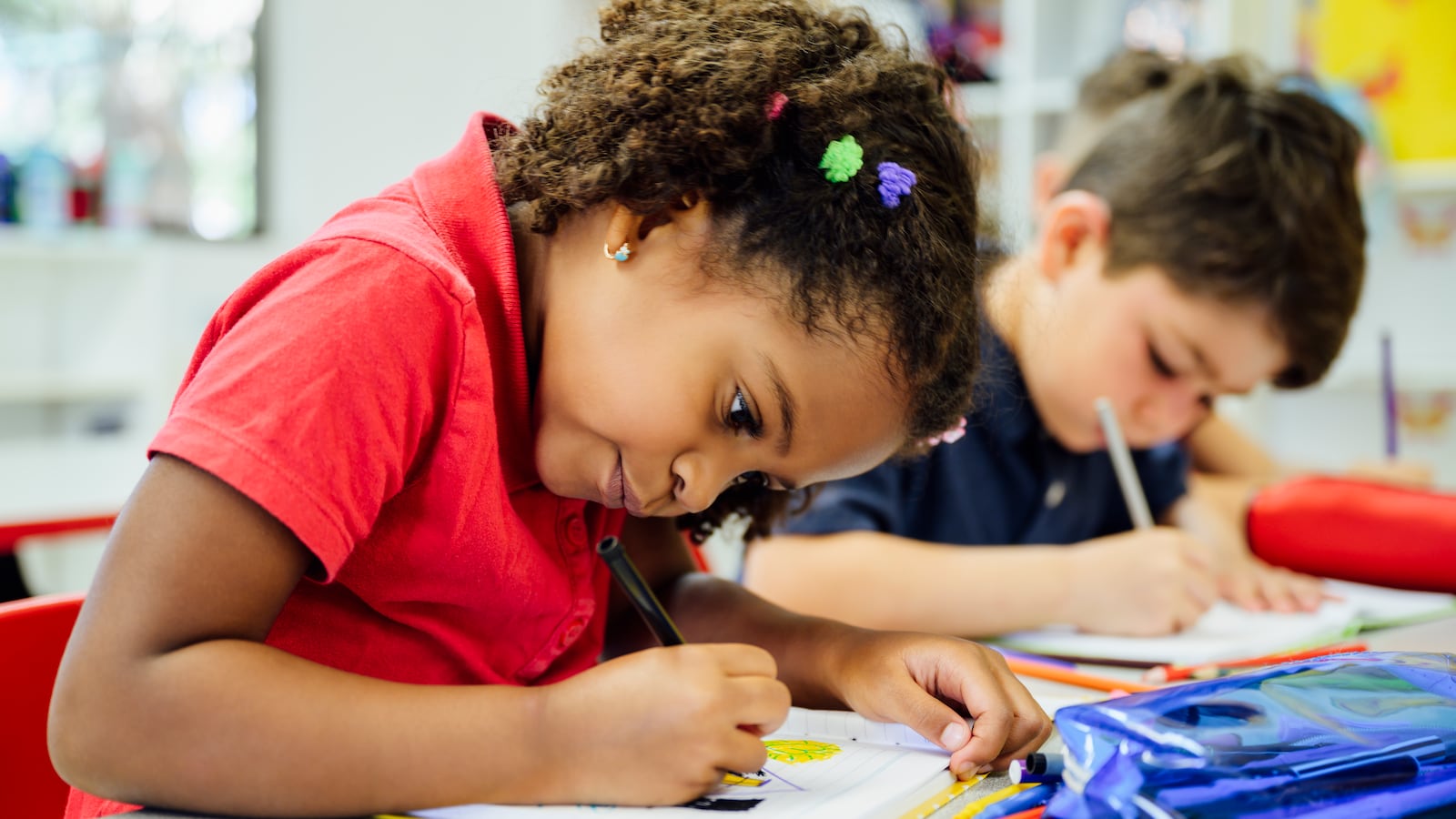 Close-up photograph of a young Black schoolgirl sitting at classroom table and concentrating on drawing with colored pencils next to a young white boy.