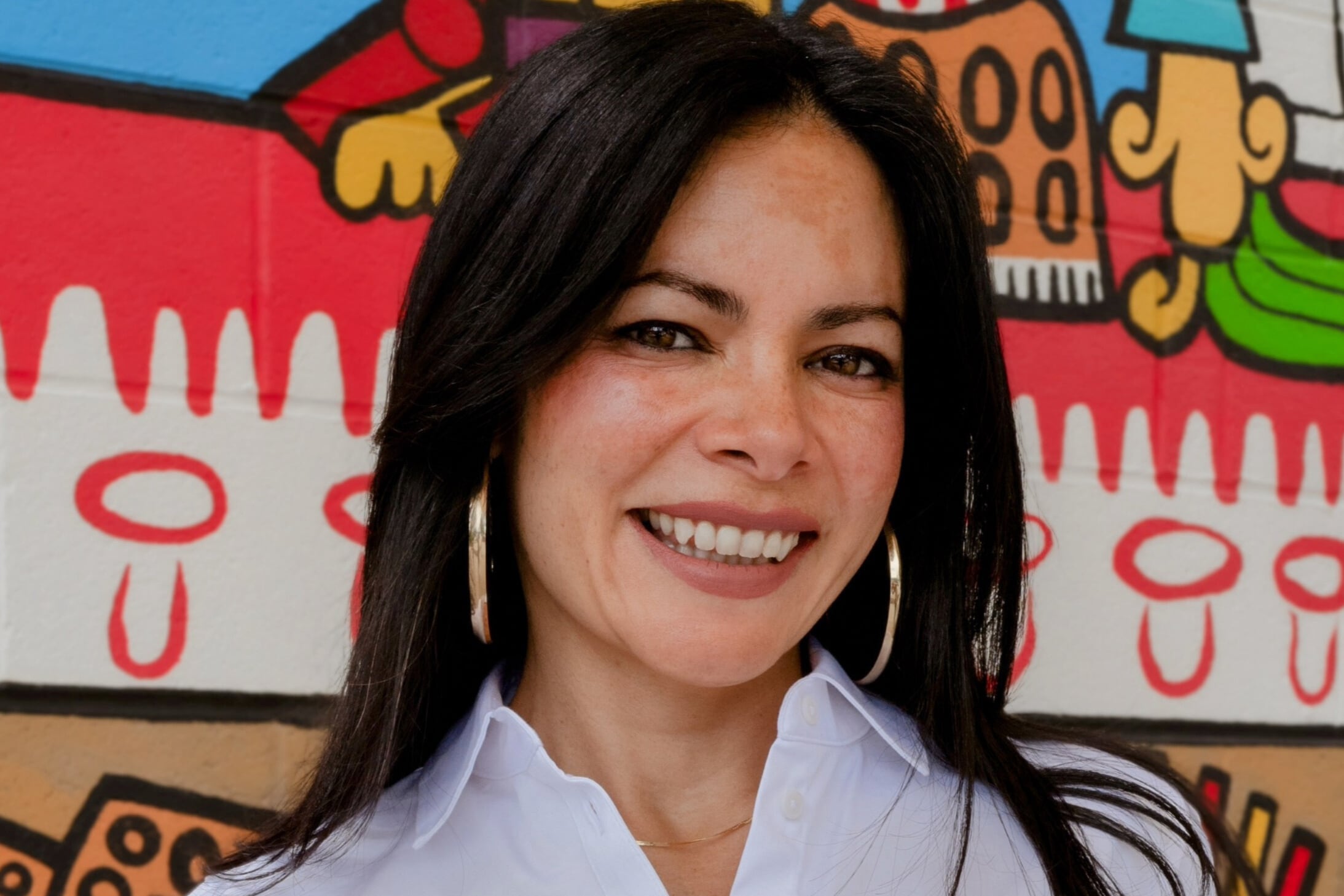 A photograph of a woman with long dark hair and wearing a white blouse, smiles for a portrait against a colorful mural in the background.