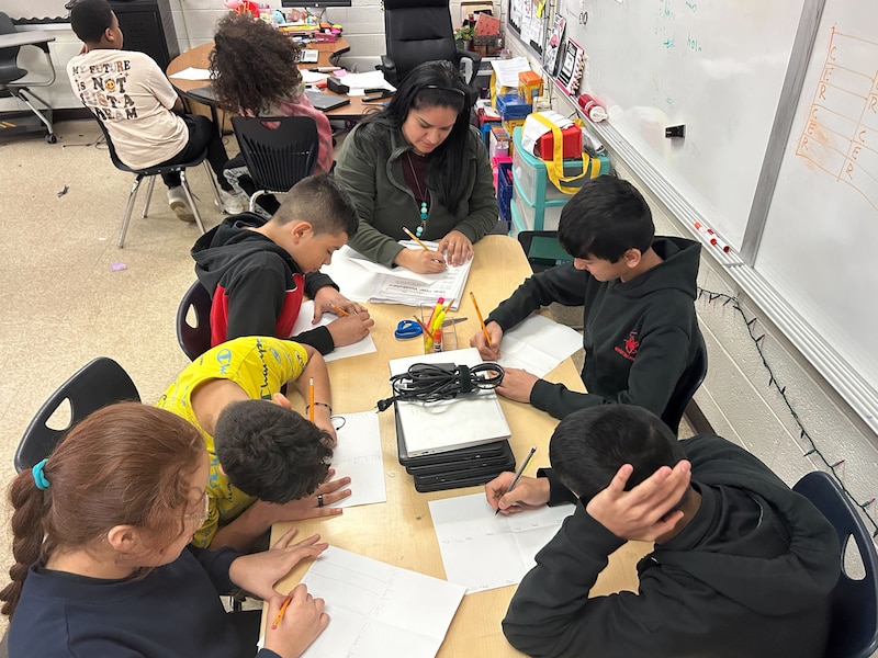 An adult with long dark hair sits at the end of a table in a classroom with five students sitting around the table while they work on class work. There are two students sitting in the background.