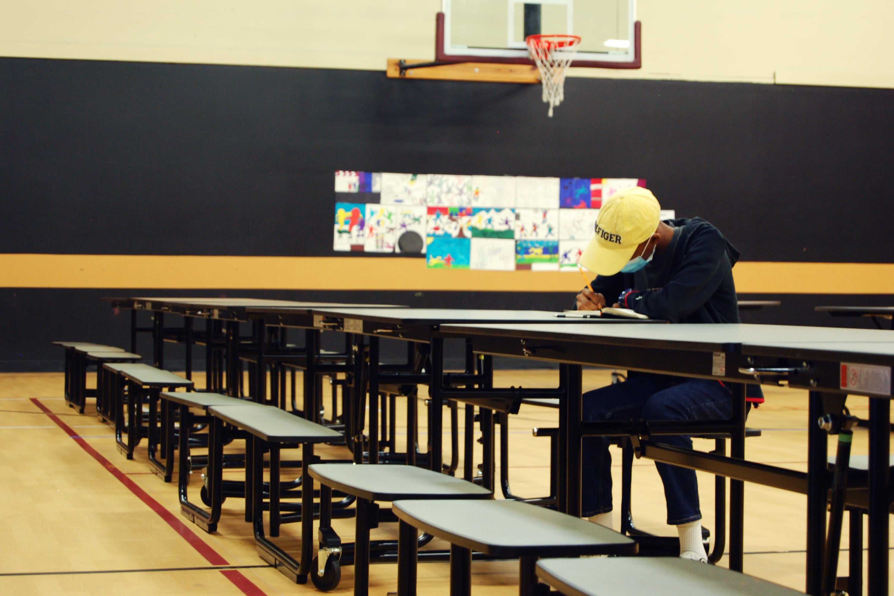 A student hearing a sweatshirt and a baseball cap sits at a cafeteria table in a gymnasium. He is writing in a notebook.