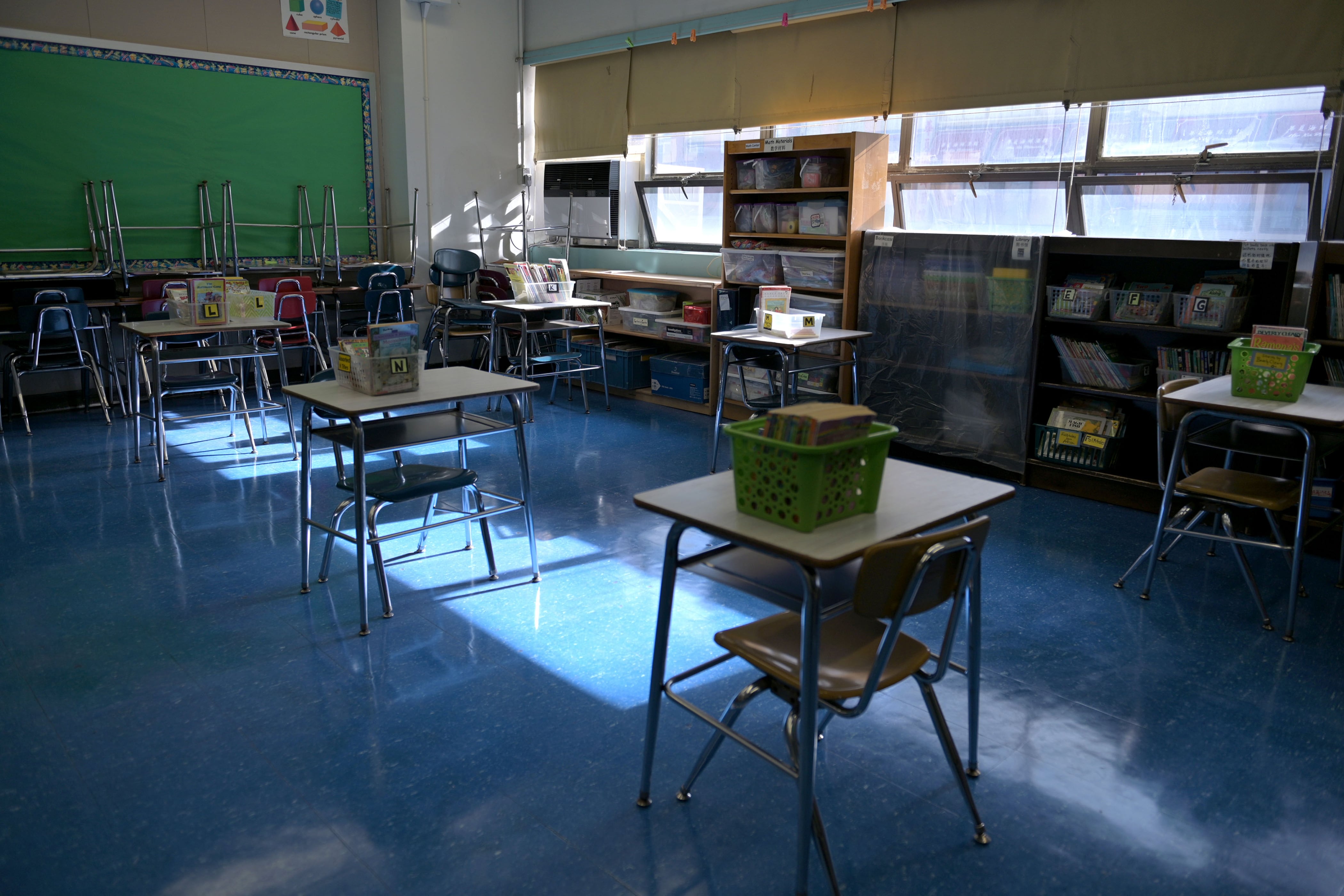 An empty classroom with the lights turned off and desks in the center of the room with a blue floor and a green chalkboard in the background.