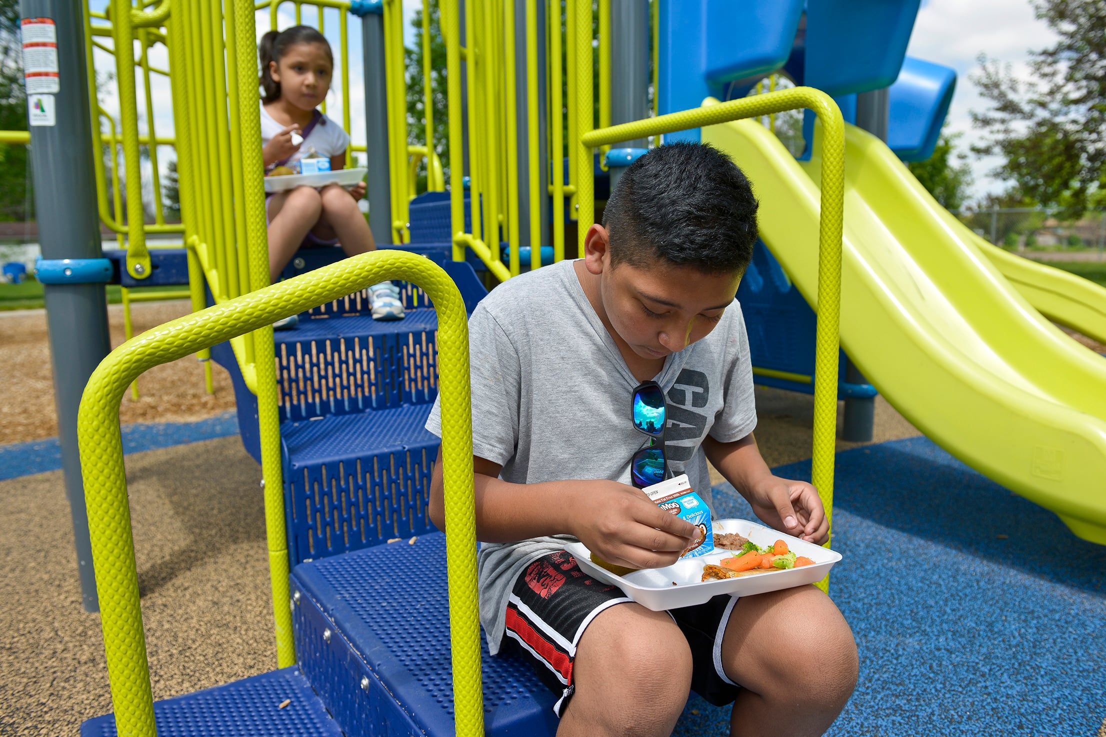 Two young students eat lunch outside on a blue and green playground.