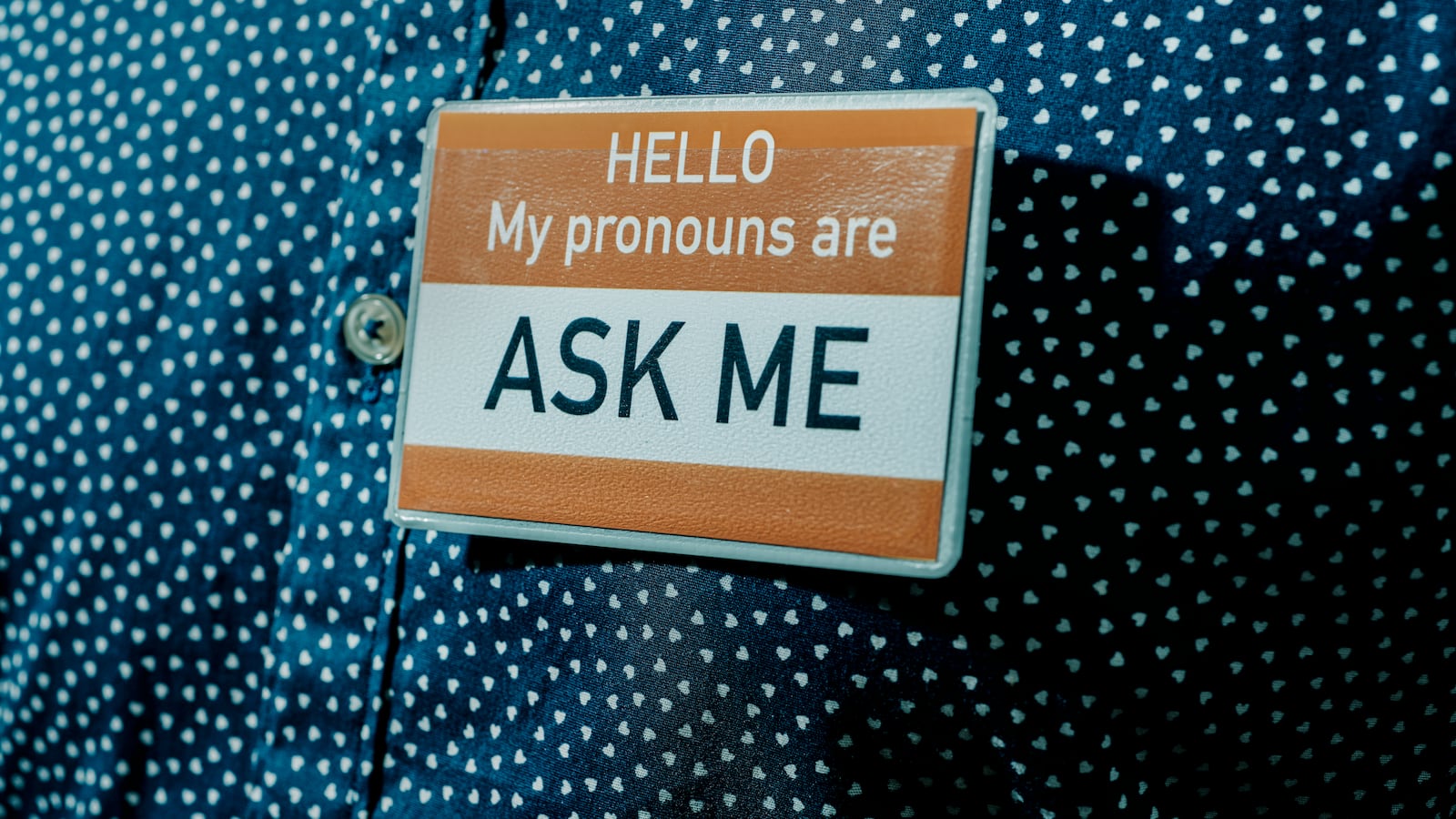 Close-up of a person wearing a blue and white shirt, and a card with the text, "Hello my pronouns are ask me" pinned in it