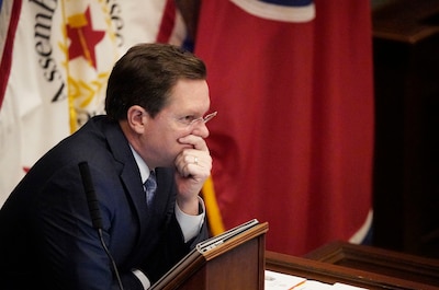 A man with brown hair and wearing a dark suit sits at a desk with flags in the back.