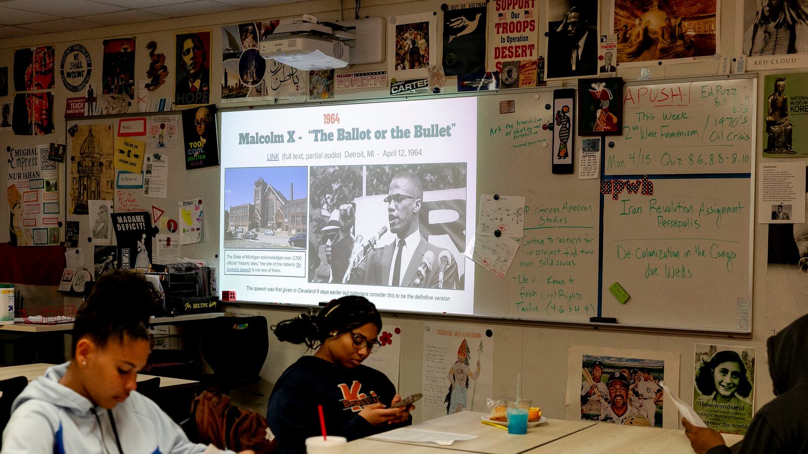 Two high school students work on classwork at desks with a large projector screen and a wall with posters in the background.