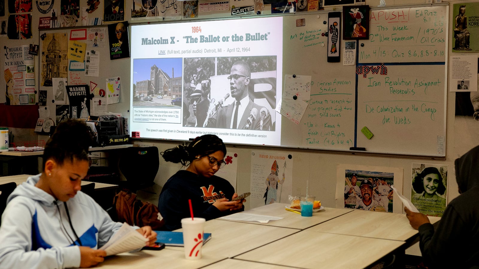 Two high school students work on classwork at desks with a large projector screen and a wall with posters in the background.