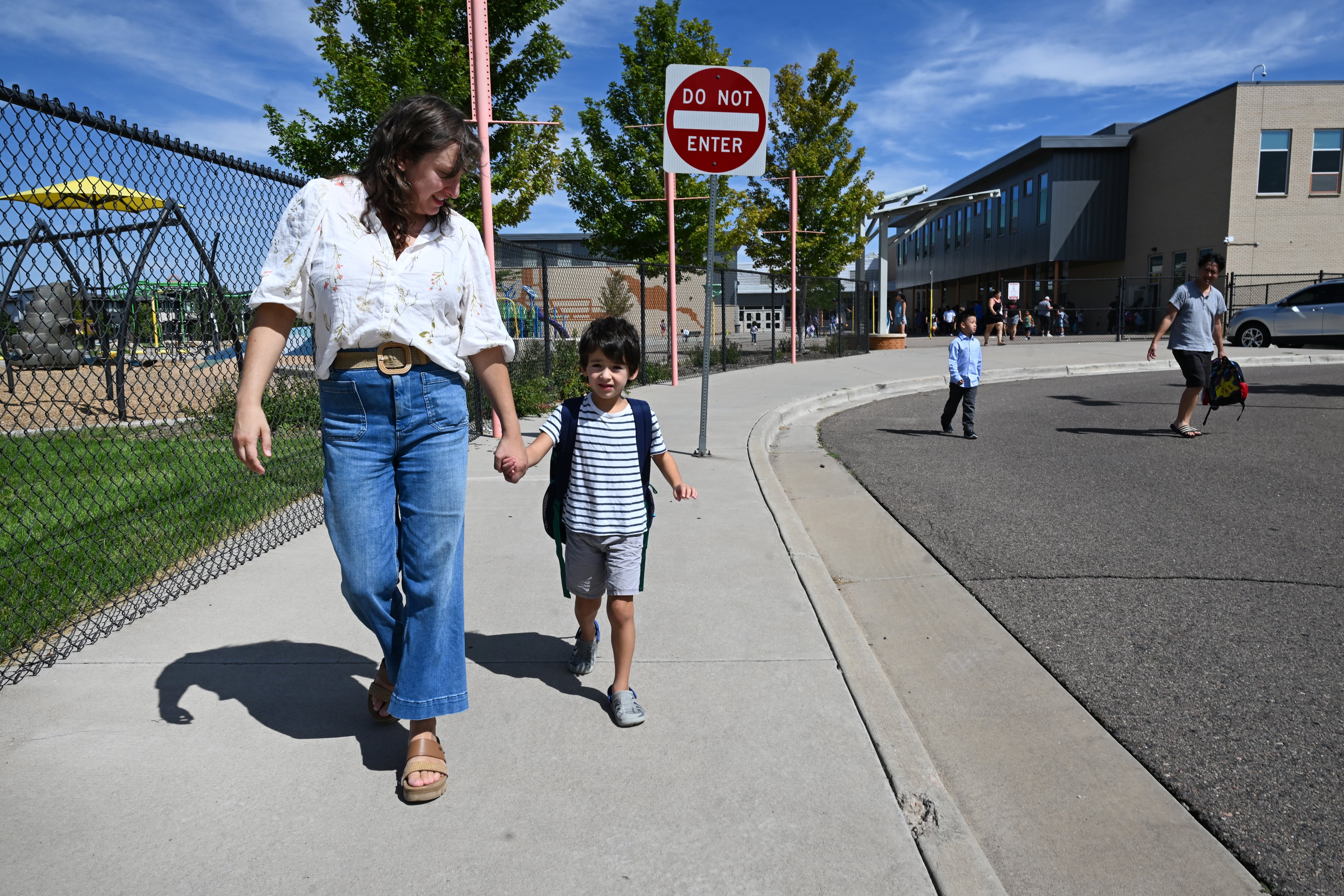 An adult woman holds the hand of a young boy while they walk away from a school building.