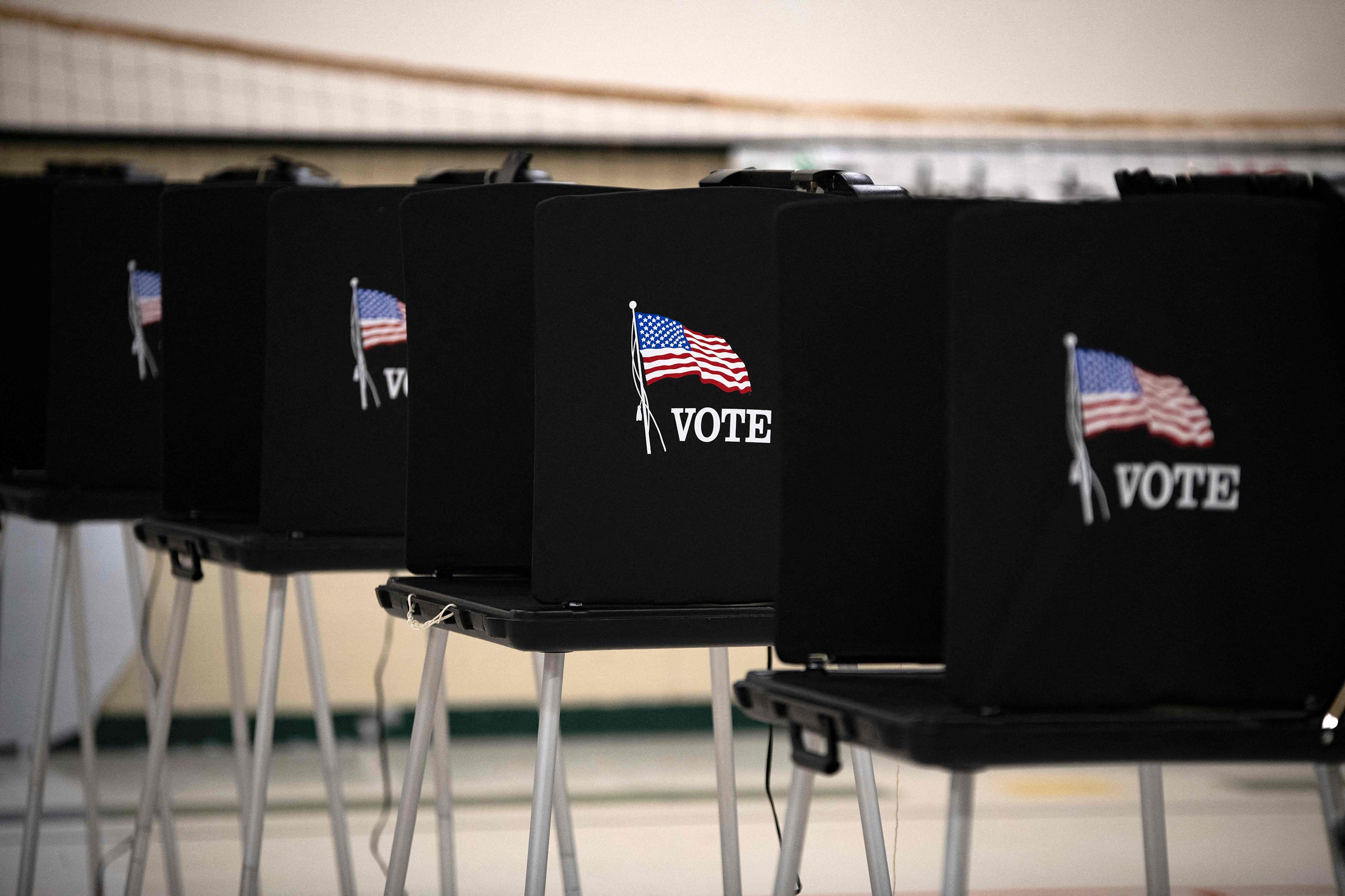 A photograph of a row of black voting booths with an American flag and the word "VOTE" on the side in an elementary school gym.