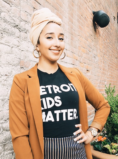 A woman wearing a tan head scarf and an orange sweater smiles at the camera standing in front of a brick wall.