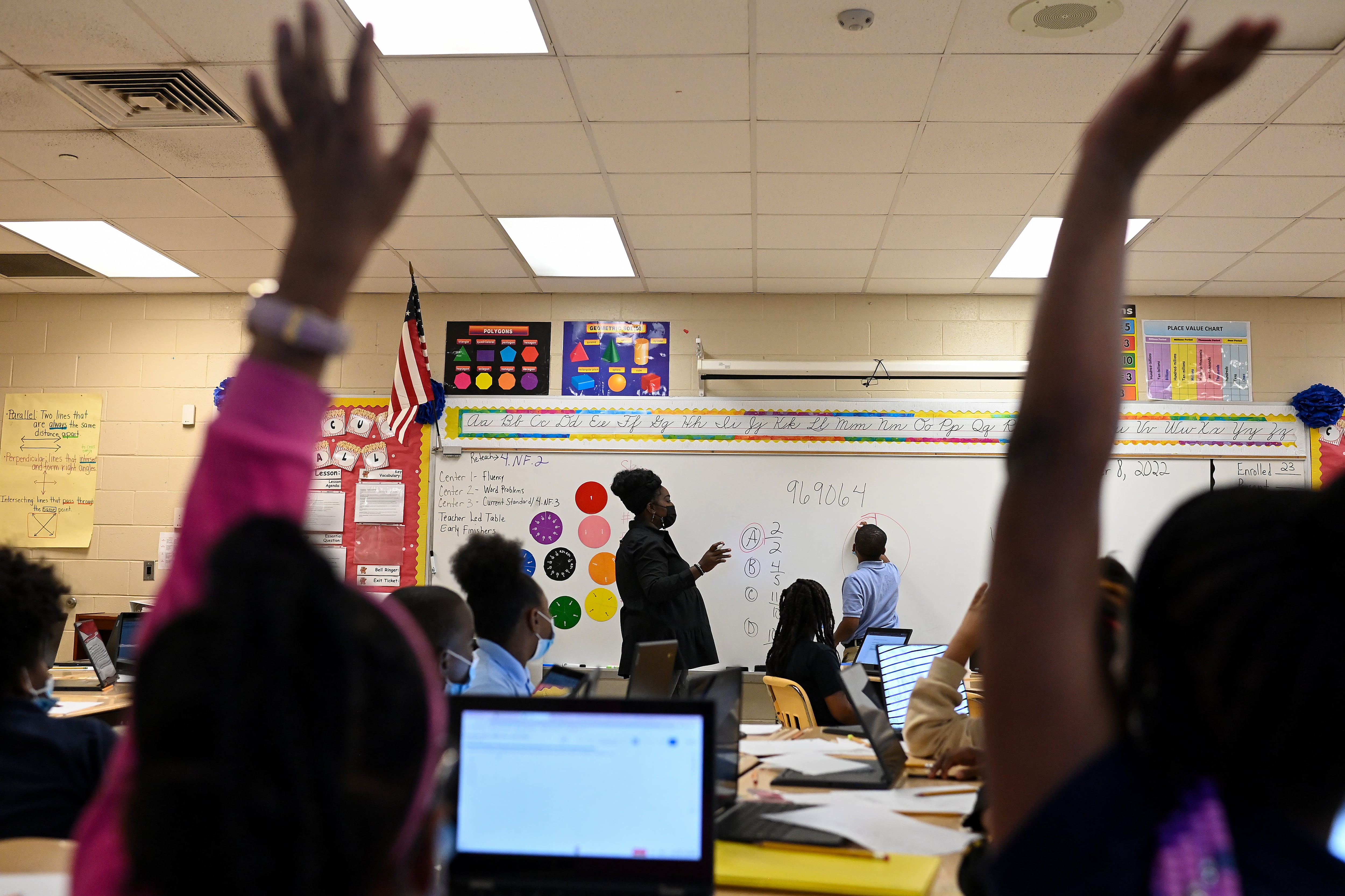 A woman in a mask stands in front of a white board with colored wheels on it as children raise their hands in front of her. 