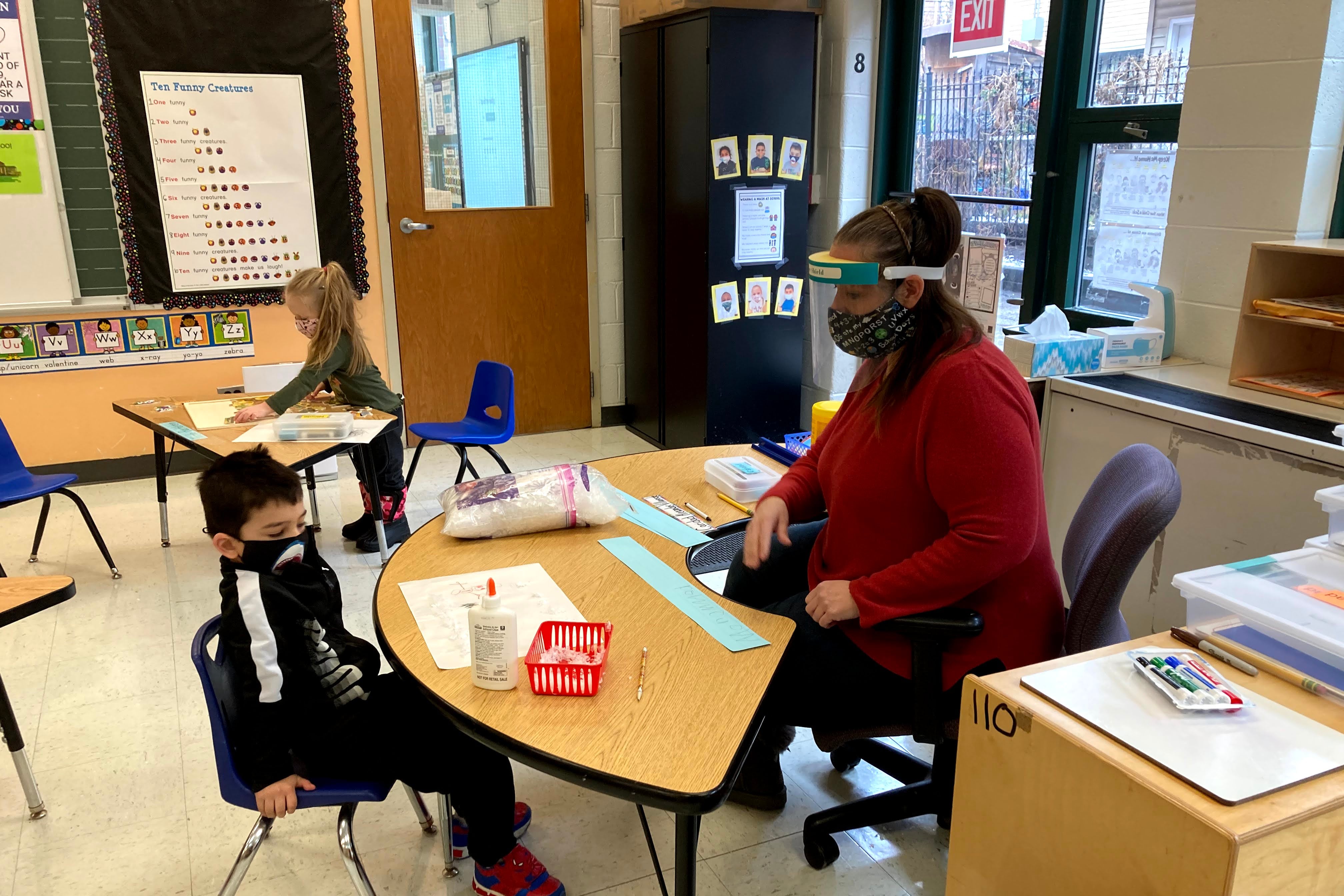 Masked teacher and masked student face each other across a semicircular desk in a classroom.
