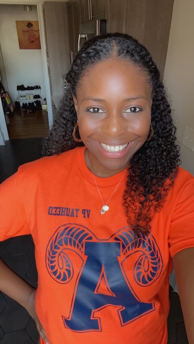 A Black woman wearing an orange t-shirt smiles at the camera.