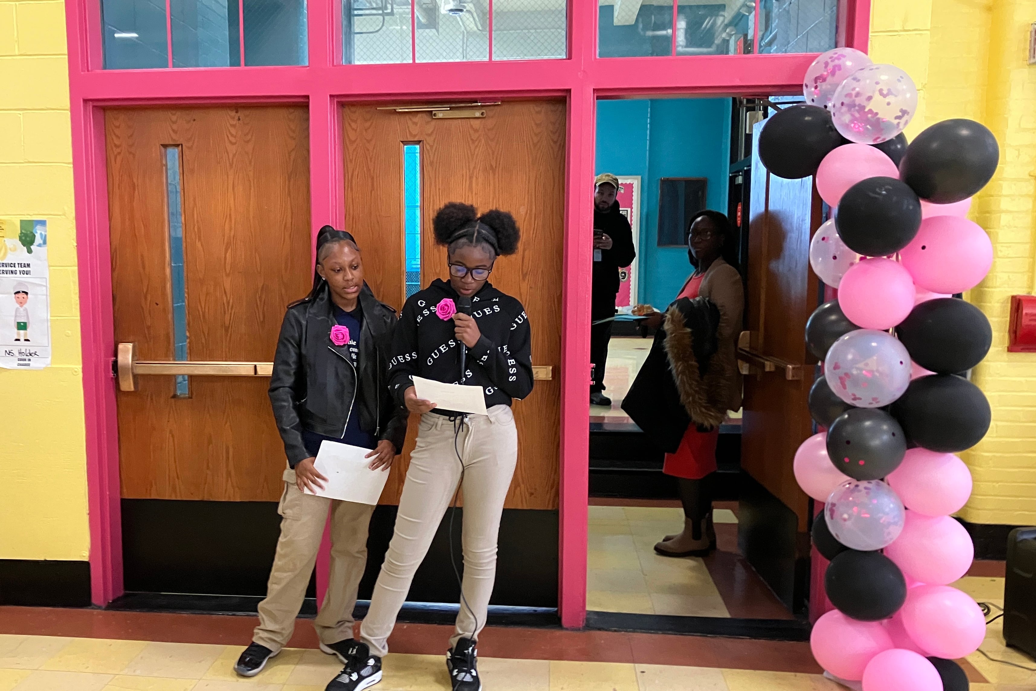 Two students stand near a doorway next to a tower of balloons in a hallway.