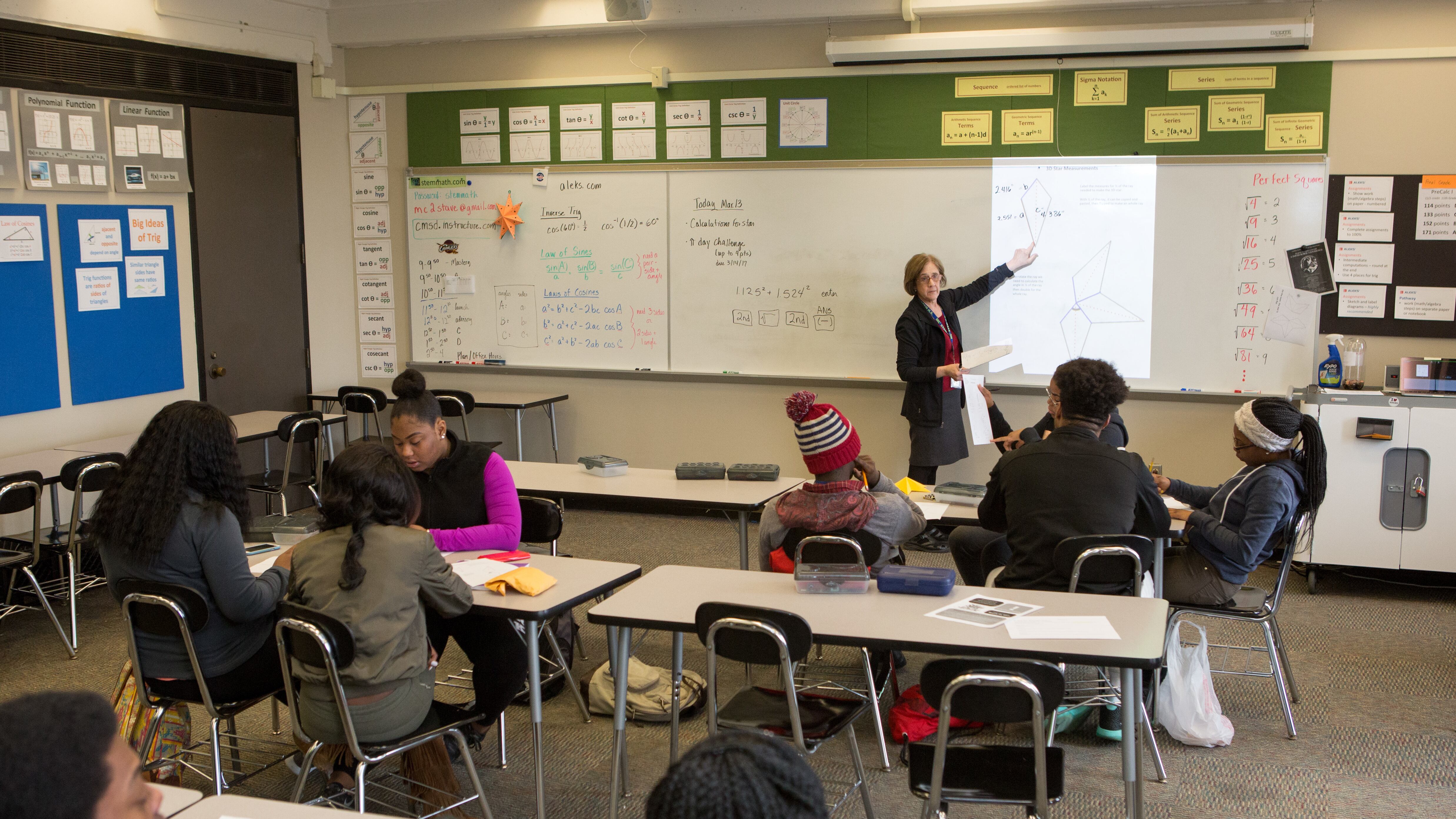 A teacher stands at the front of a classroom pointing at a white board as three rows of students look on.