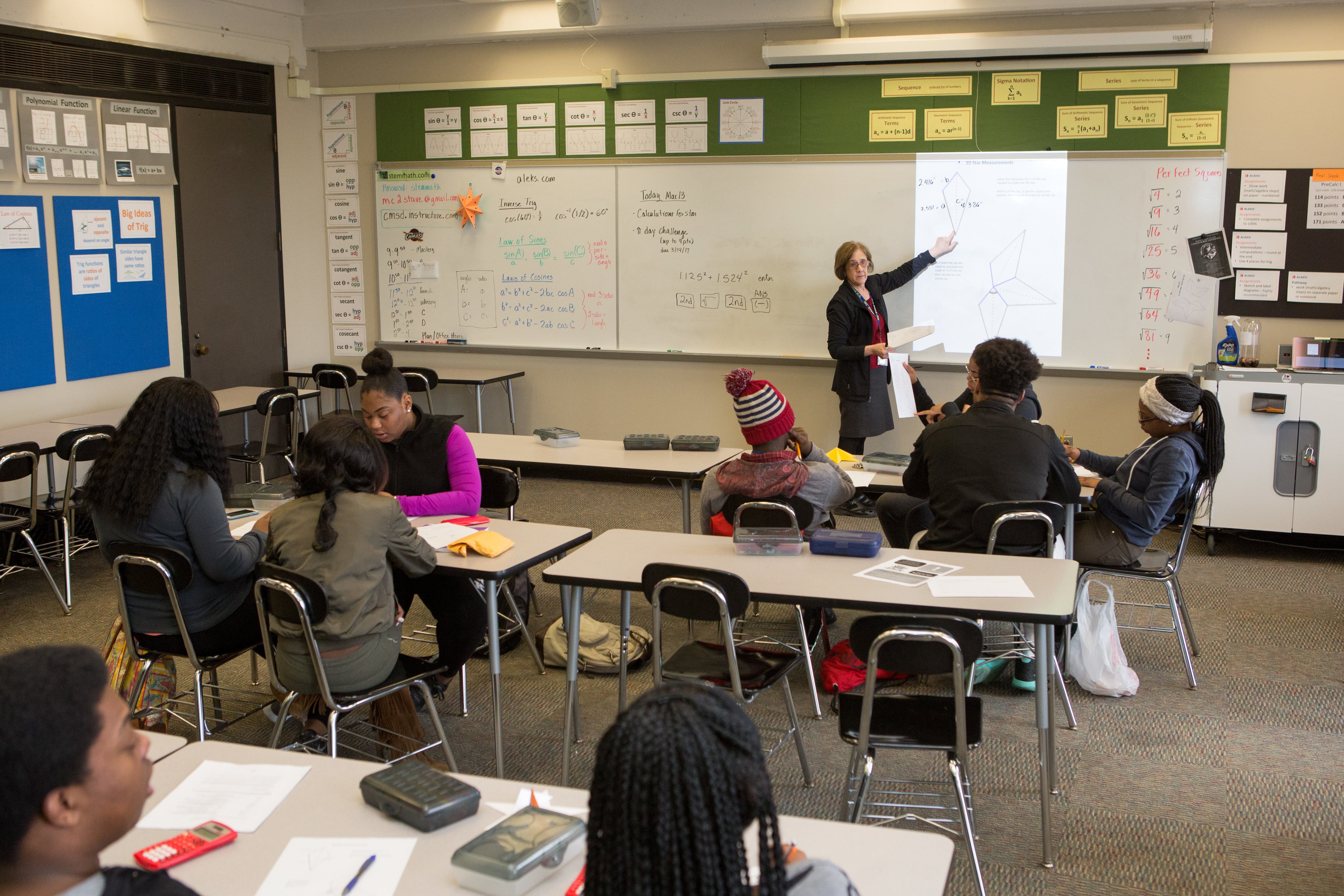 A teacher stands at the front of a classroom pointing at a white board as three rows of students look on.
