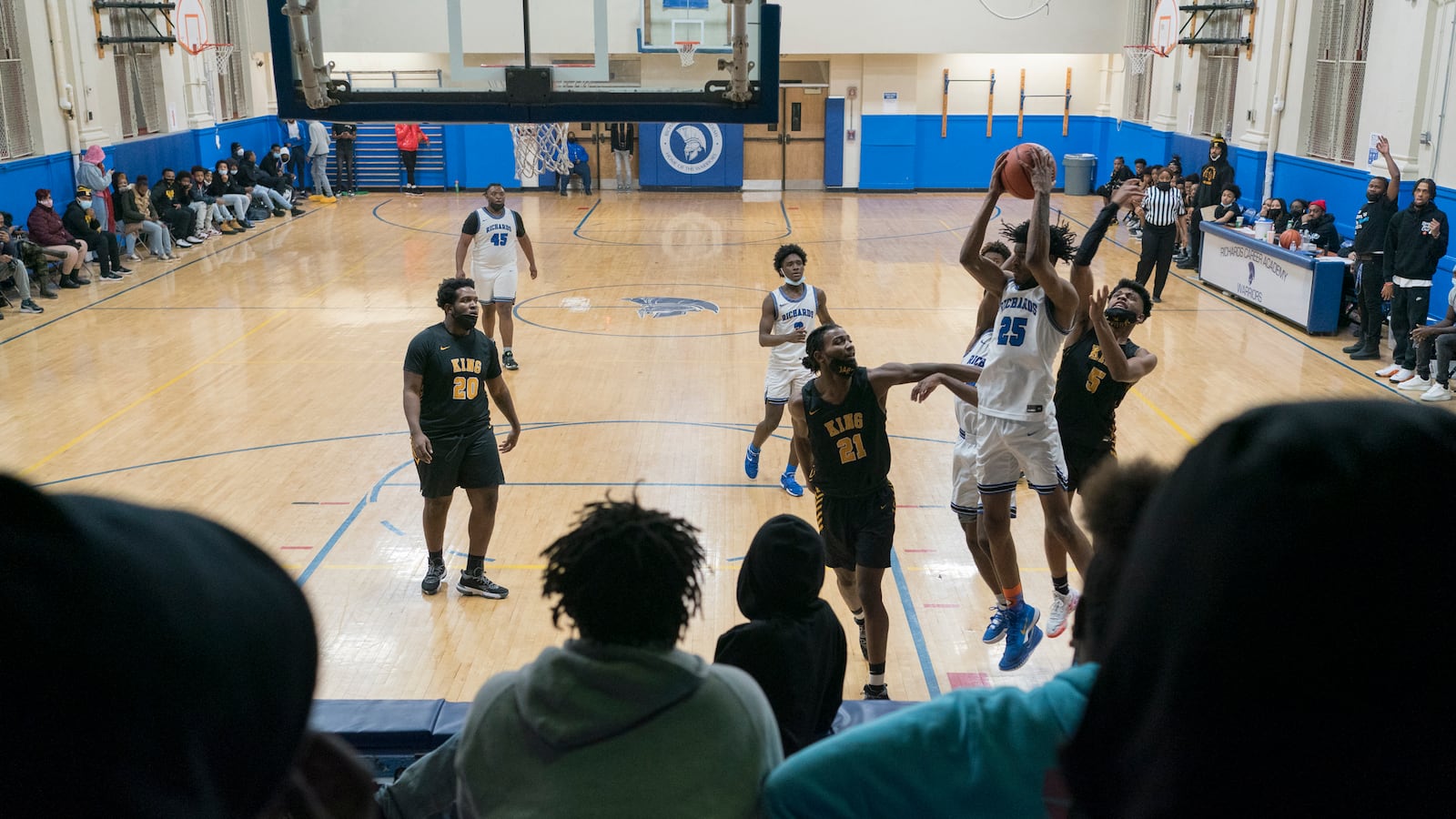 Spectators watch the action of a high school basketball game from the bleachers, as the players on the court jump for a rebound.