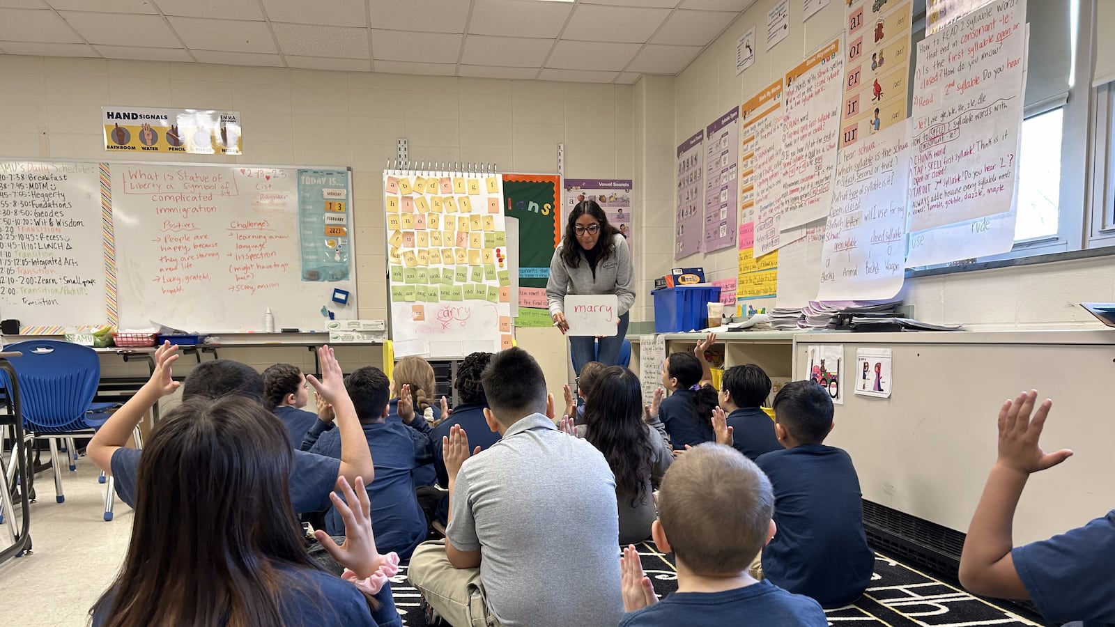 A teacher holds a small white board that says “marry” in front of a class of roughly a dozen students sitting on a carpet.