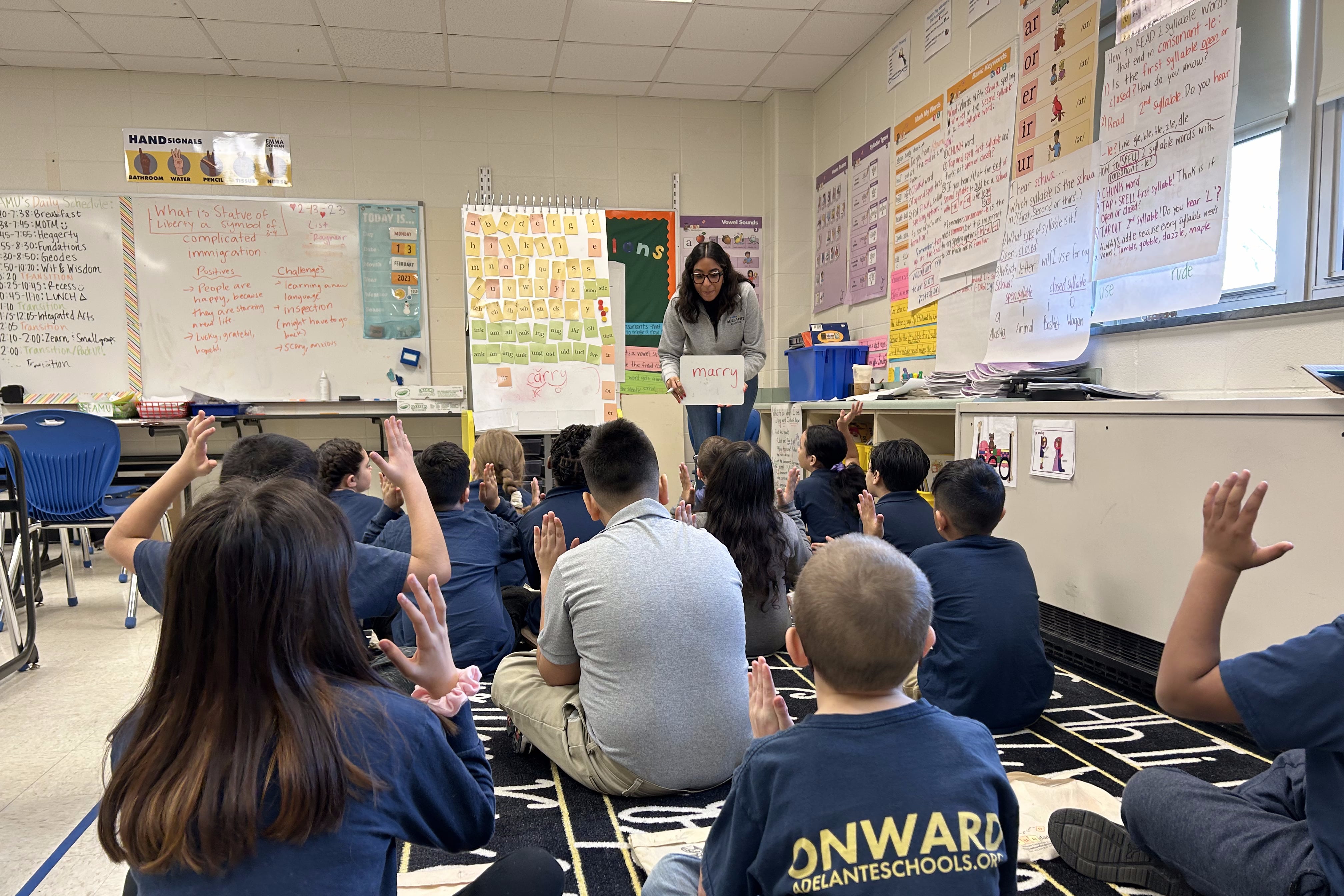 A teacher holds a small white board that says “marry” in front of a class of roughly a dozen students sitting on a carpet.