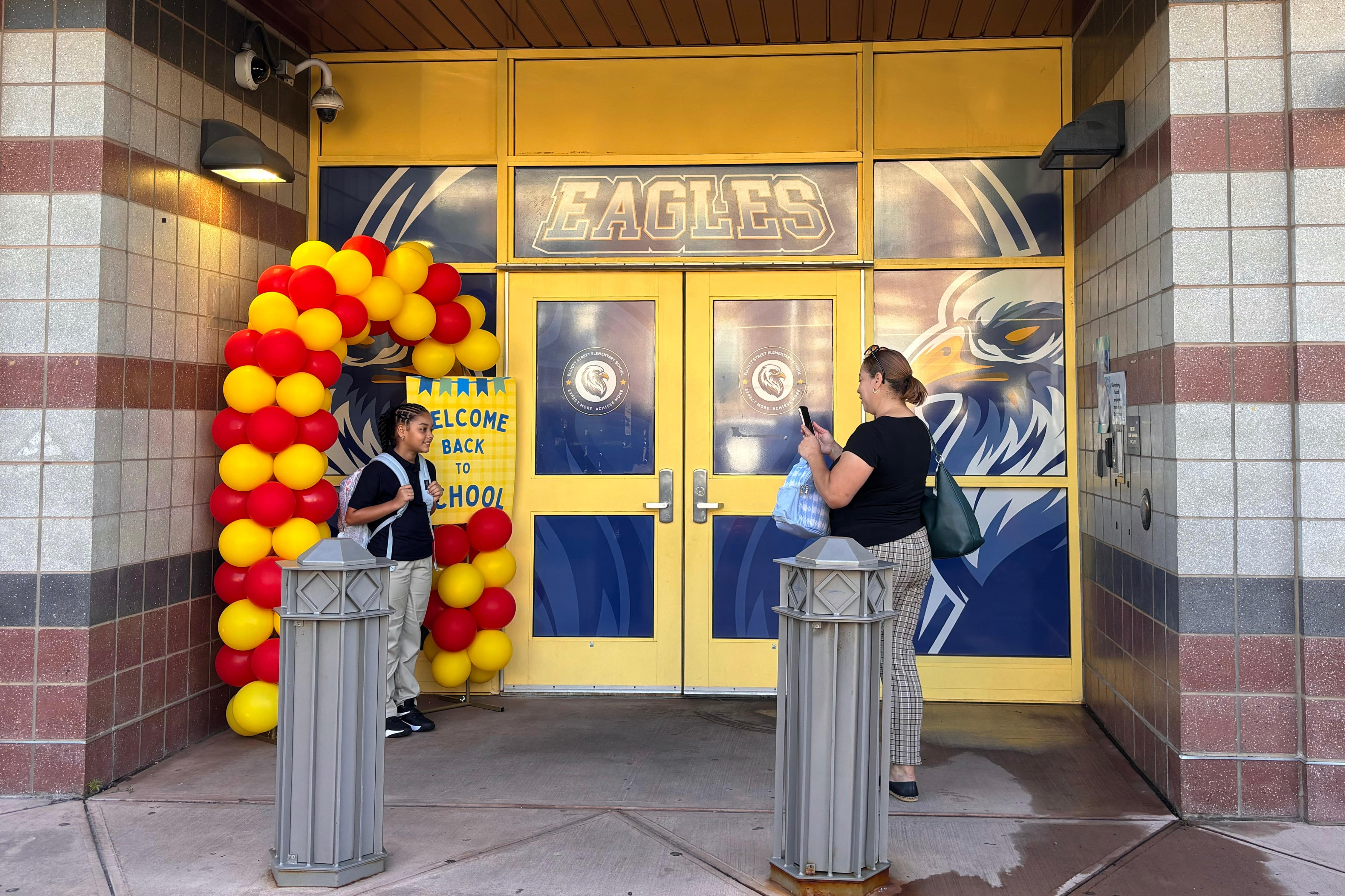 An adult woman makes a cellphone photo of a young student under a balloon arch in front of a school entrance.