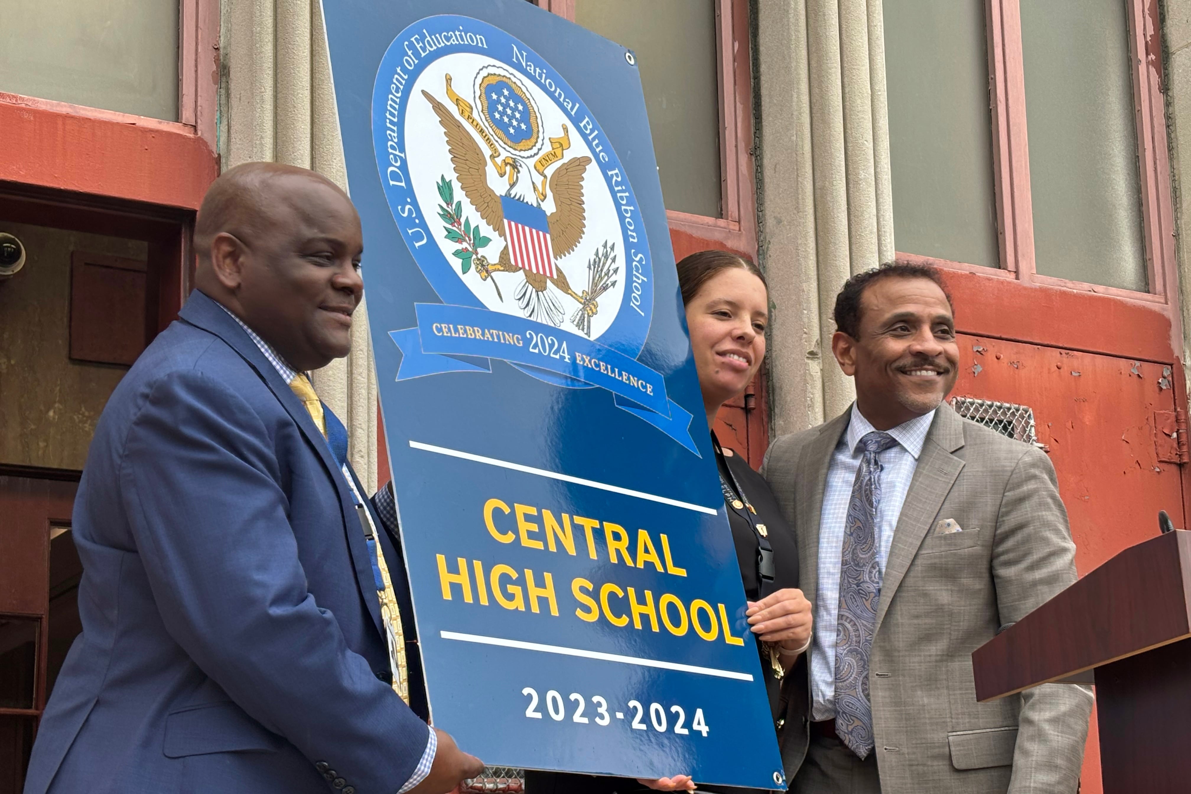 Three people stand holding a large banner in front of a red wall.