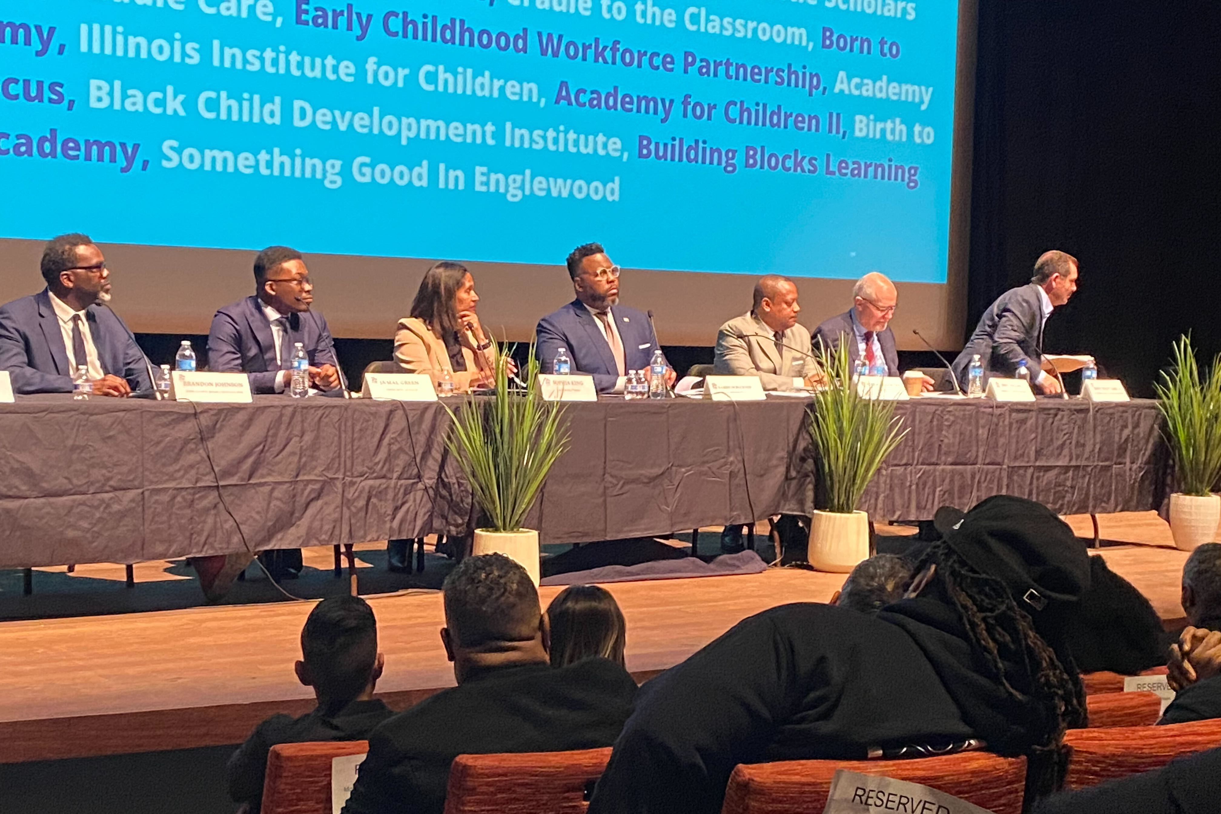 Seven of the eight mayoral candidates in Chicago sat on stage at table covered in Black with green plants in front. In the foreground of the photo people sit in red chairs.