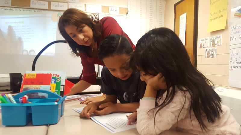 A teacher stands over two young students at their desk, and watches as they practice reading together from the same book.