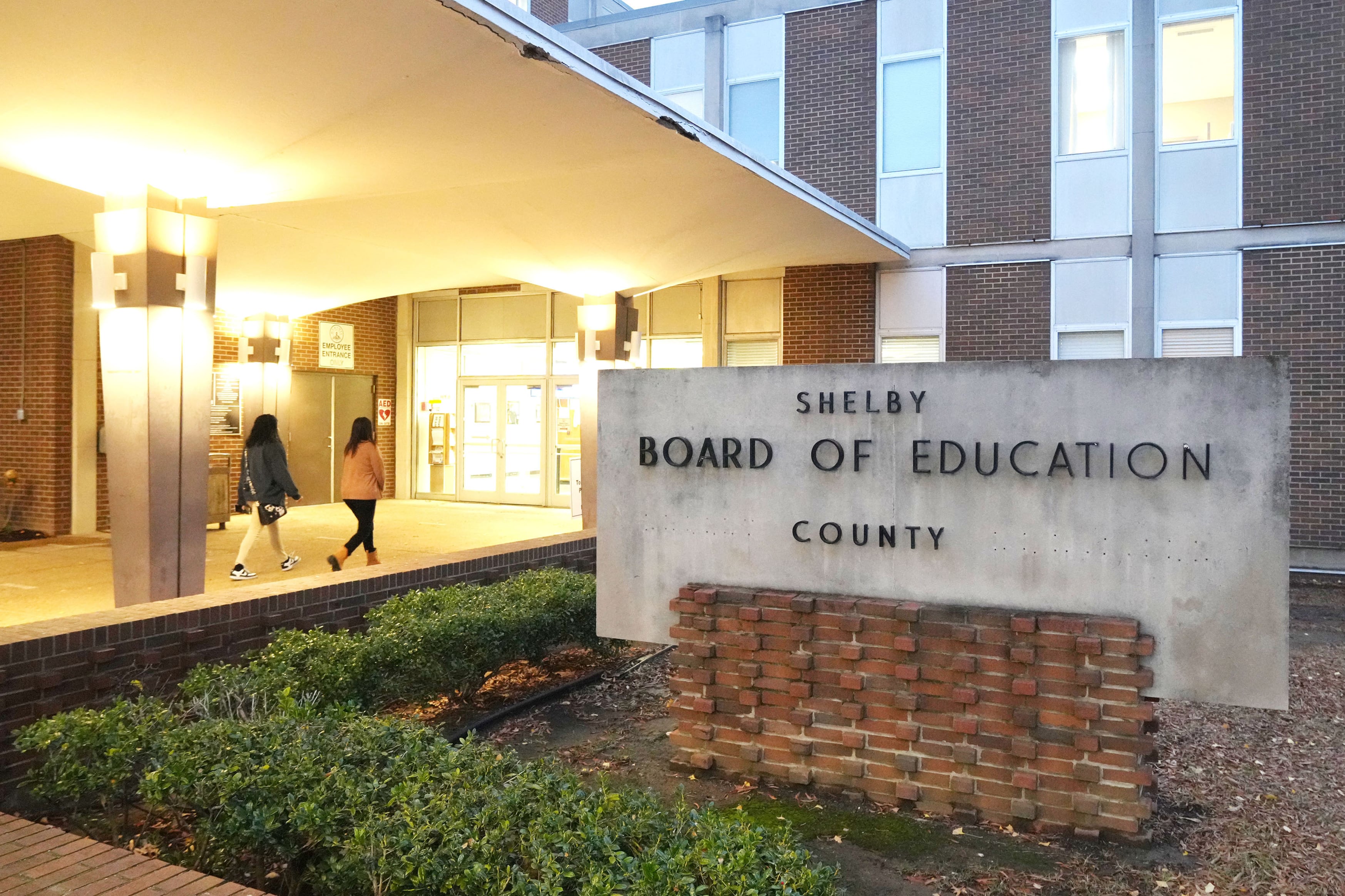 A photograph of two people walking into a large building. There's a sign on the side that reads "Shelby County Board of Education."