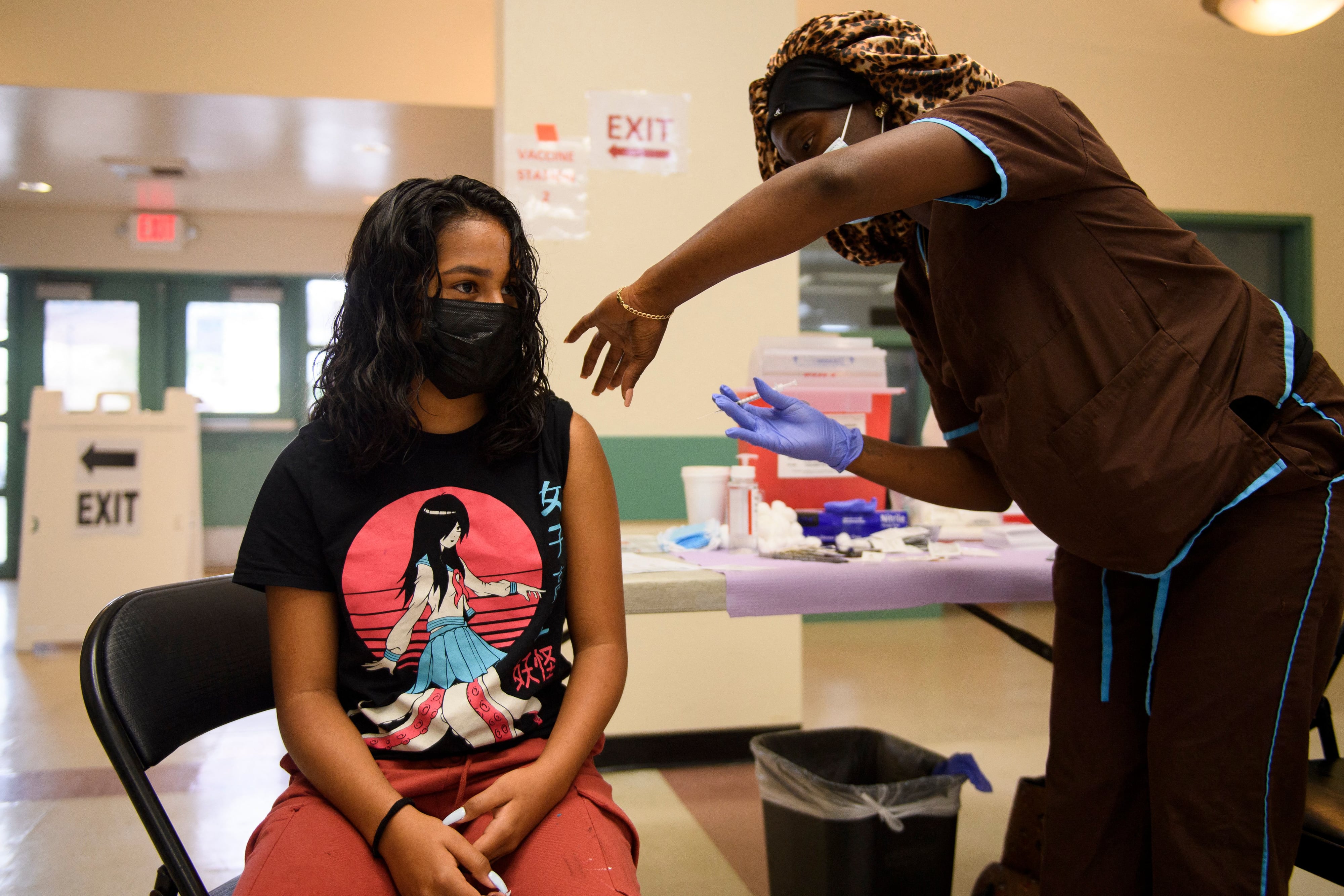 A young woman wearing a black and red japanese art-inspired shirt receives a dose of the COVID vaccine from a nurse.