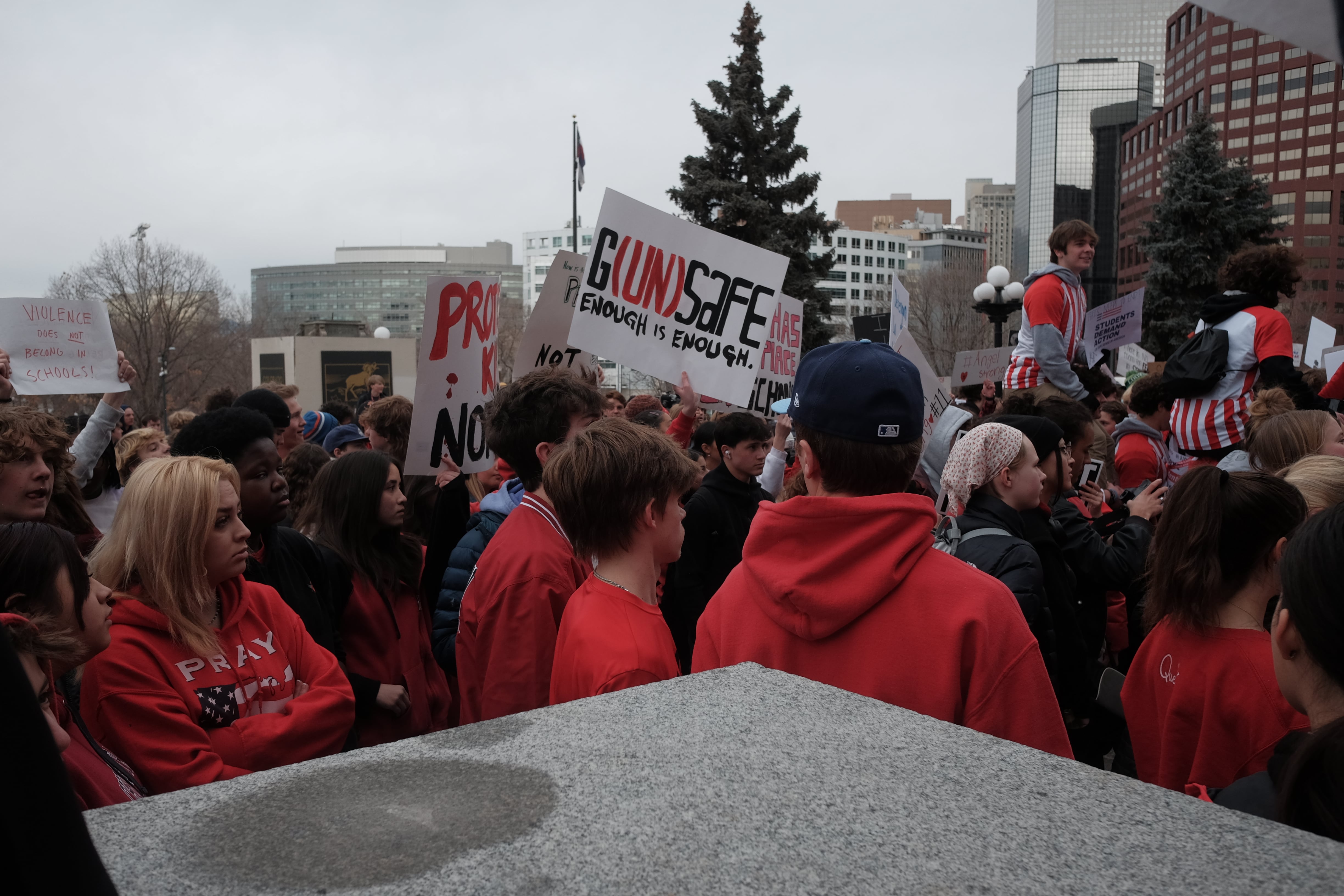 High school students dressed in red gather in front of the Colorado State Capitol. One sign emerges from the crowd reading “G(UN)SAFE Enough is Enough.” Downtown office buildings can be seen in the background.