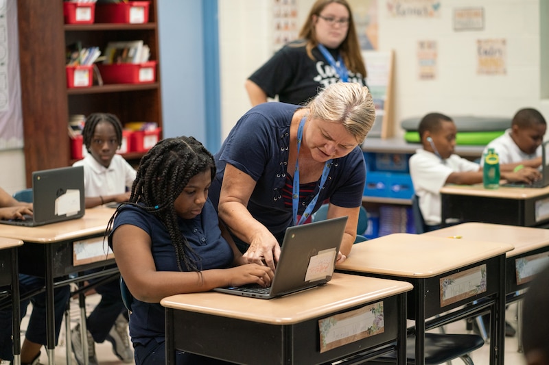 An adult wearing a dark blue shirt stands next to a student with long dark hair and wearing a blue shirt in a classroom full of people in the background.