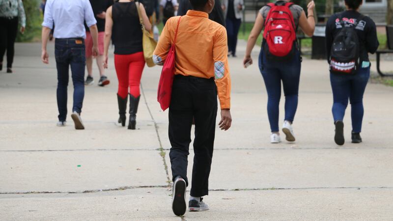 A young man wearing an orange shirt, black pants and carrying a red bag walks on a college campus behind other college students.
