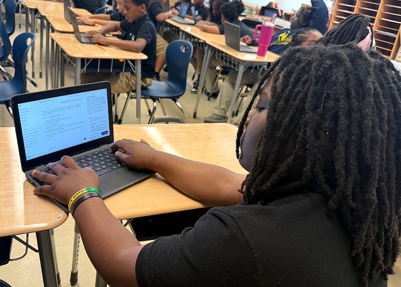 A student in a dark t-shirt works on a laptop in a classroom.