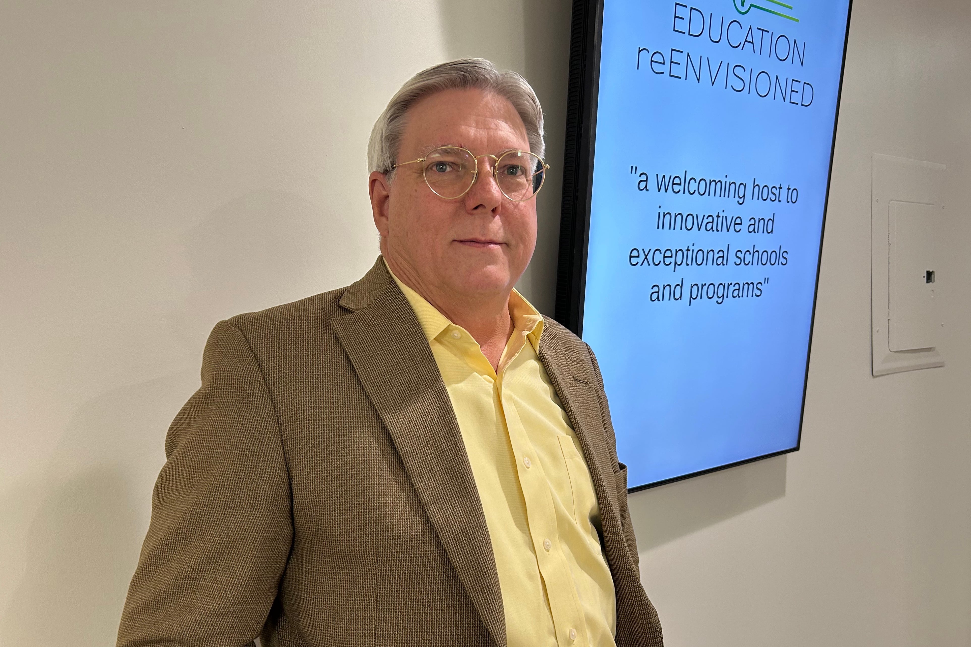 A photograph of a white man in a brown suit and glasses posing for a photograph in front of a white wall with a large TV screen.